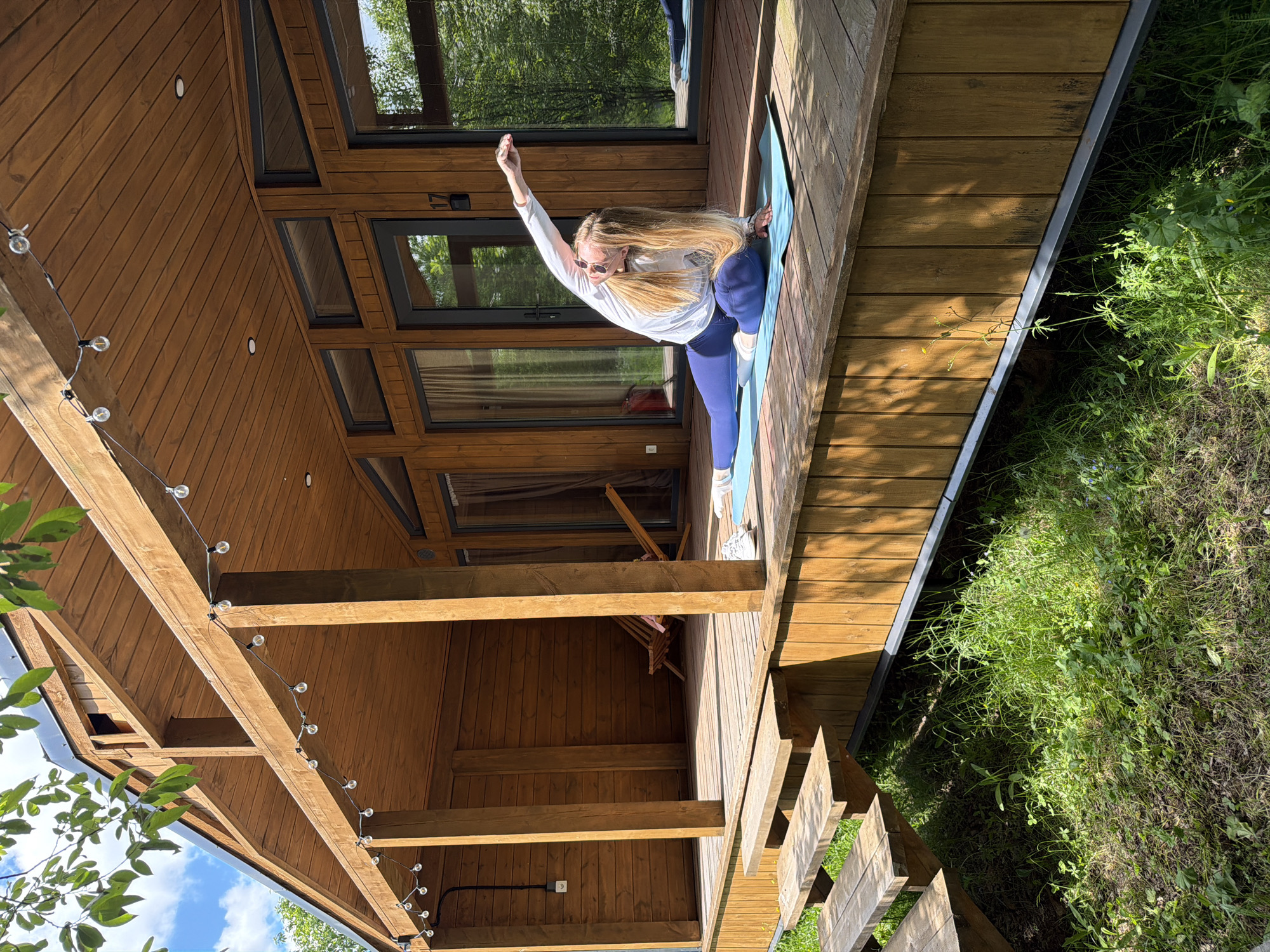 A woman with long blonde hair, wearing sunglasses, a white long-sleeved top, and blue yoga pants, is performing a yoga pose on a blue mat. She is on a wooden deck next to a modern wooden house, with her right arm extended upwards and her left hand near her left foot, executing a stretching posture akin to Extended Side Angle Pose.
The setting is outdoors on a sunny day, with strong shadows cast by the wooden structures, suggesting either morning or late afternoon. The house is constructed from warm-toned wooden planks and features numerous large windows with dark frames, reflecting the surrounding green trees and the blue sky. String lights with circular bulbs are strung along the overhead wooden beams of the deck, which also has a wooden staircase leading down to a natural grassy area with lush green vegetation. Inside the house, visible through one of the windows, is a wooden chair. The number "17" is visible on the wooden frame above a window on the house's exterior. The scene portrays a calm moment of exercise in a tranquil, nature-integrated environment.