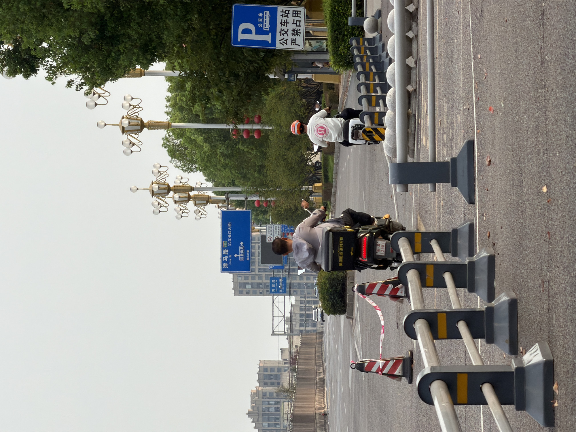 The image depicts a street scene in Jiangjin, China. Two individuals on scooters are visible, traveling in the same direction. The scooter in the foreground is ridden by a person wearing a light-colored hoodie, while the scooter further back is ridden by a person wearing a white t-shirt with a red and white helmet. Both scooters have storage boxes attached to the rear.
The street is paved and appears to be a road with multiple lanes. Several traffic cones, some marked with red and white stripes, are present, suggesting some form of roadwork or temporary lane closure. A series of dark gray barriers with horizontal silver rails and yellow accents line the right side of the road, separating it from a grassy area with trees.
Road signs are visible in the background. One prominent blue sign indicates directions with Chinese characters and the route number "653". Another blue sign displays a "P" symbol indicating parking, along with Chinese text that translates to "Bus Stop" and "No Parking". Decorative streetlights with multiple globes are also visible along the roadside. In the distance, tall buildings and other structures suggest an urban environment. The sky is overcast and a pale white. The overall lighting suggests it is daytime, possibly late afternoon or early morning given the diffused light.