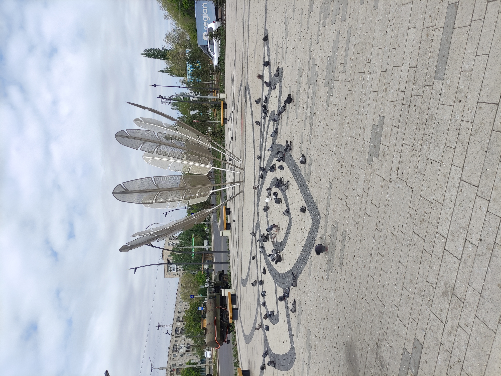 The image depicts a public space in Pavlodar, Kazakhstan. A prominent sculpture of metal feathers stands on a paved surface. The pavement features a dark-grey stylized floral design. Numerous pigeons are scattered around the pavement, concentrated within the floral design. In the background, a road and building are visible. A utility truck is parked next to the road. A white bus and other vehicles can also be seen in the background. The sky is overcast.