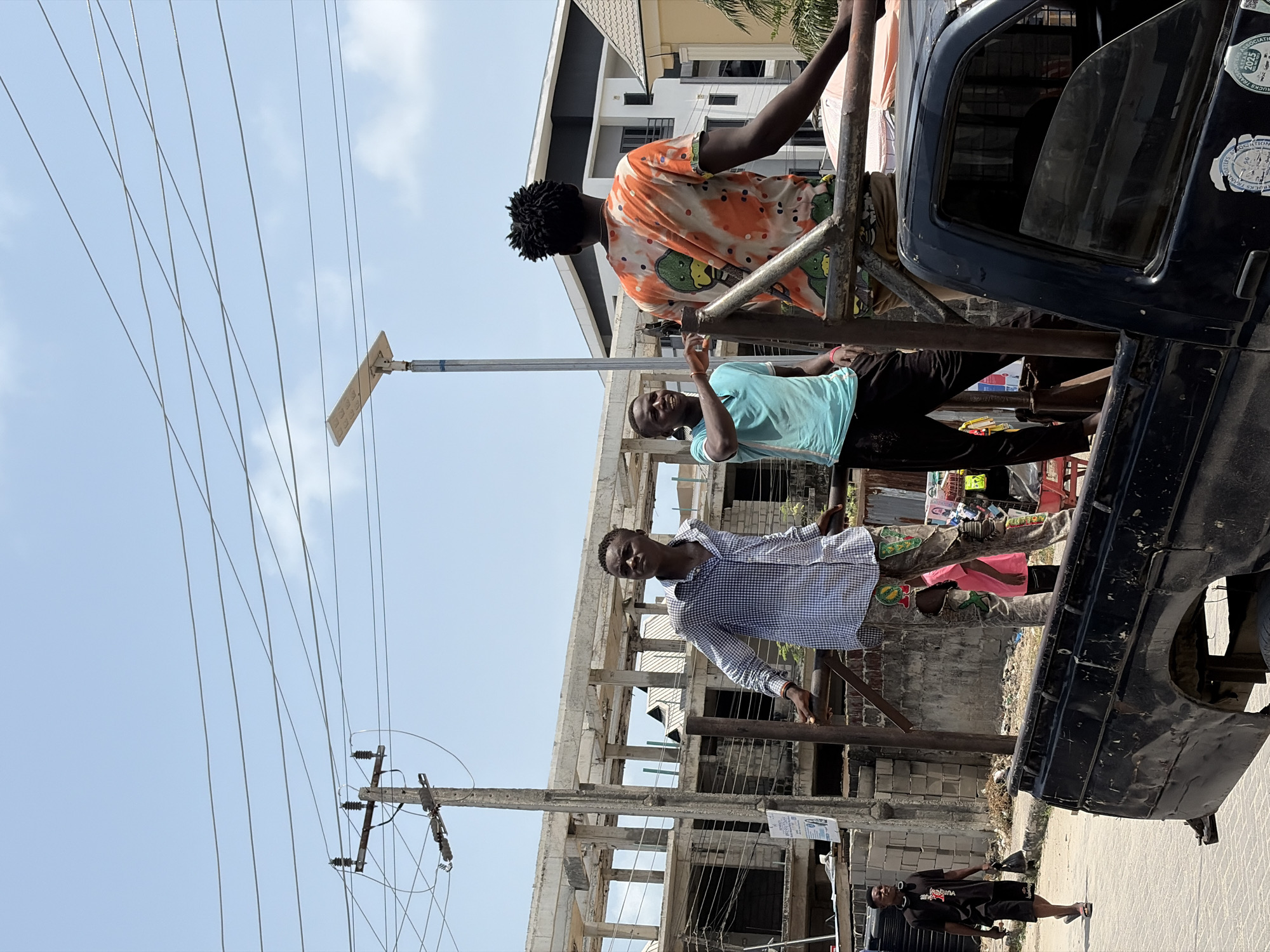 This image captures a bright daytime scene in Igboefon, Nigeria, featuring a group of men by a truck amidst a developing urban landscape. The sky is clear blue with sparse clouds, indicating sunny weather.
Four men are visible. Three men are positioned in the back of a dark-colored truck, which appears to be a utility or pickup truck, possibly modified with a metal frame. The leftmost man, wearing an orange and white patterned shirt, stands with his back to the camera, looking towards ongoing construction. Next to him, a man in a light blue t-shirt faces the camera, smiling and raising his right hand in a friendly gesture. The third man in the truck, closest to the camera, wears a blue and white checkered shirt and looks directly at the viewer with a neutral expression. On the ground to the right, a fourth man in a black t-shirt and shorts stands, looking towards the truck. He holds an object in his left hand.
The setting includes buildings under various stages of construction, with exposed concrete frames prominent in the background. A more complete building with a dark roof is also visible further back. Numerous overhead power lines crisscross the sky, connected to a utility pole on the left. A street light equipped with what appears to be a solar panel extends into the frame from the left.
Notable details include several stickers on the truck's driver-side door; one partially legible sticker reads "ASSOCIATION OF" and "TRUCKS TRADERS," with "2023" also visible on another sticker. The ground shows some scattered debris and construction materials. The overall activity suggests a casual gathering or form of transport, common in the region.