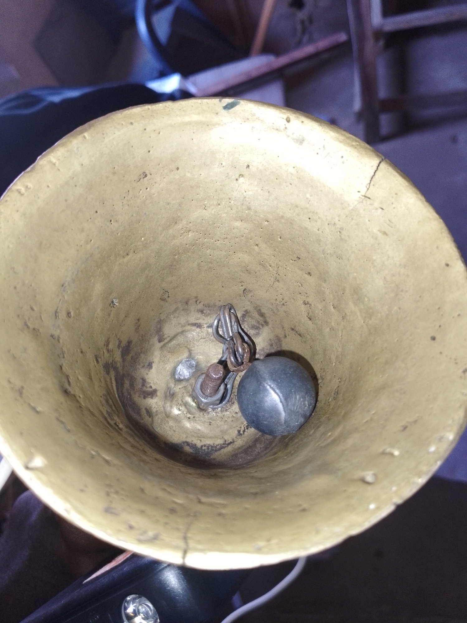 The image shows the inside of a brass bell. A metal clapper, attached to a chain, hangs from the top of the bell. The bell has a rough, textured surface with some dark discoloration and small cracks. The background is out of focus but appears to be an indoor setting with furniture and possibly a window. There is no visible text. The image appears to be a close-up, still shot.