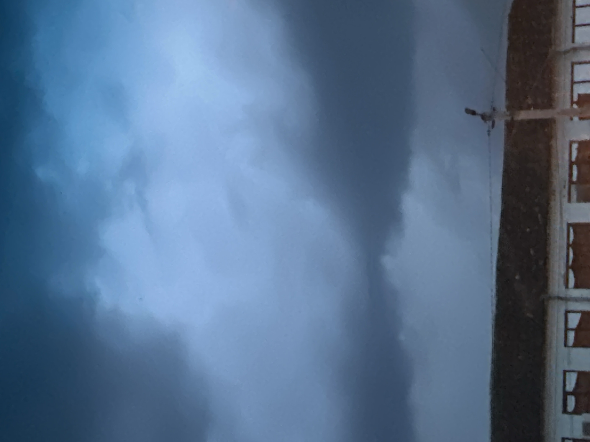 The image shows a cloudy sky over a building in Shatava, Ukraine. The sky is a deep blue with dark, heavy clouds that appear to be gathering. The building below has a dark roof and white walls with multiple windows. A utility pole with wires and what appears to be a light fixture is visible in the foreground. The overall impression is of an overcast day, possibly before or during a storm.