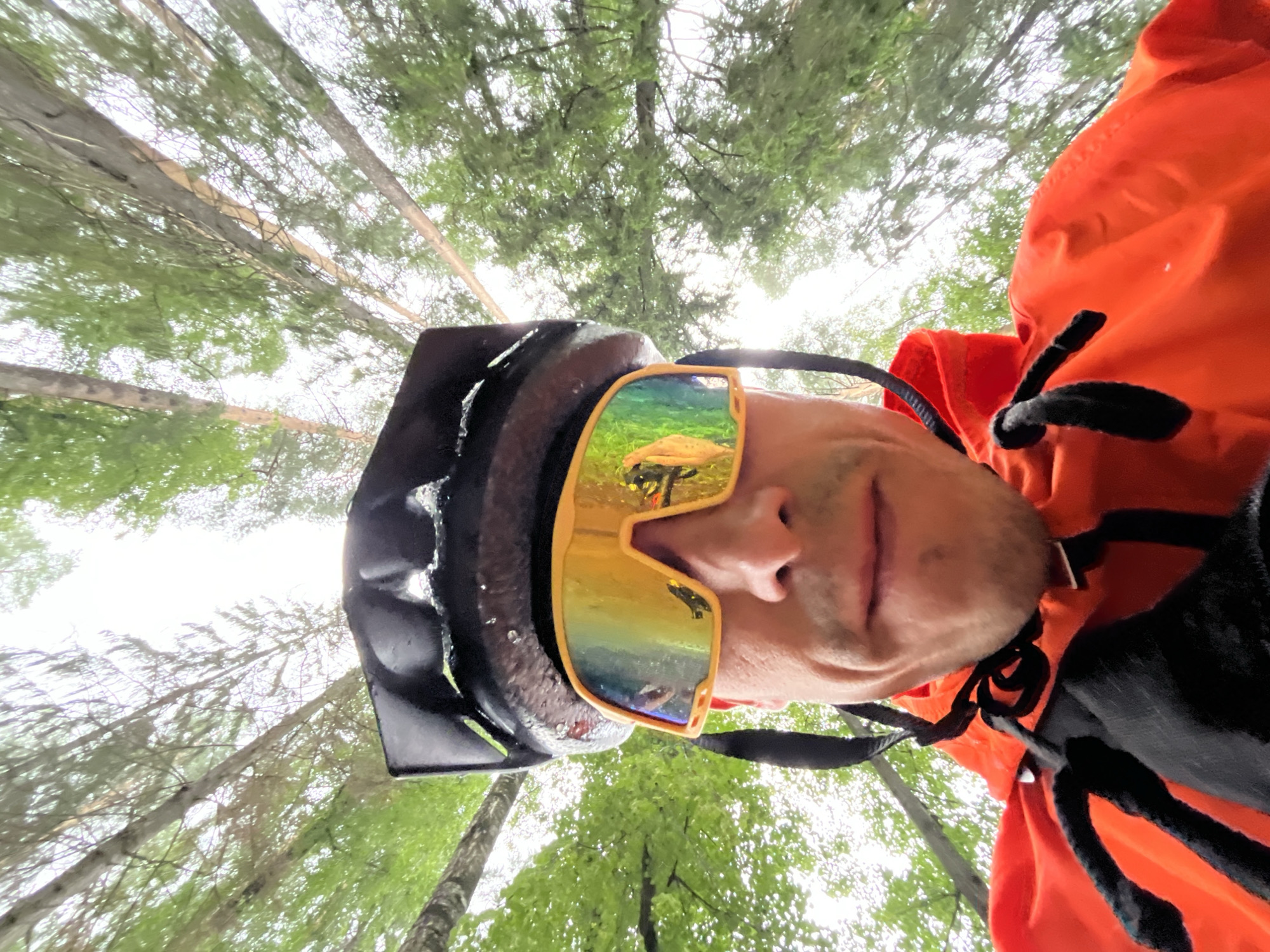 This is a low-angle, close-up, selfie-style shot of a person wearing a black helmet and vibrant orange outerwear, likely a jacket or vest, against a backdrop of a forest canopy. The subject's face is partially visible, with the focus on their mouth and nose, and they are wearing reflective sunglasses with a rainbow-like tint. The sunglasses reflect an image of green grass and a yellow object, possibly a mushroom or a piece of equipment.
The scene is set within a forest, with tall, thin trees dominating the view. The perspective is from below, looking upwards through the branches and leaves towards a bright, overcast sky. The overall impression is that of an outdoor activity, possibly cycling or hiking, given the helmet and activewear. The weather appears to be slightly damp, as indicated by water droplets visible on the helmet. The lighting suggests it is daytime, likely overcast, contributing to the diffused light in the scene. The location is identified as Chaikovsky, Russia. There is no visible text in the image.