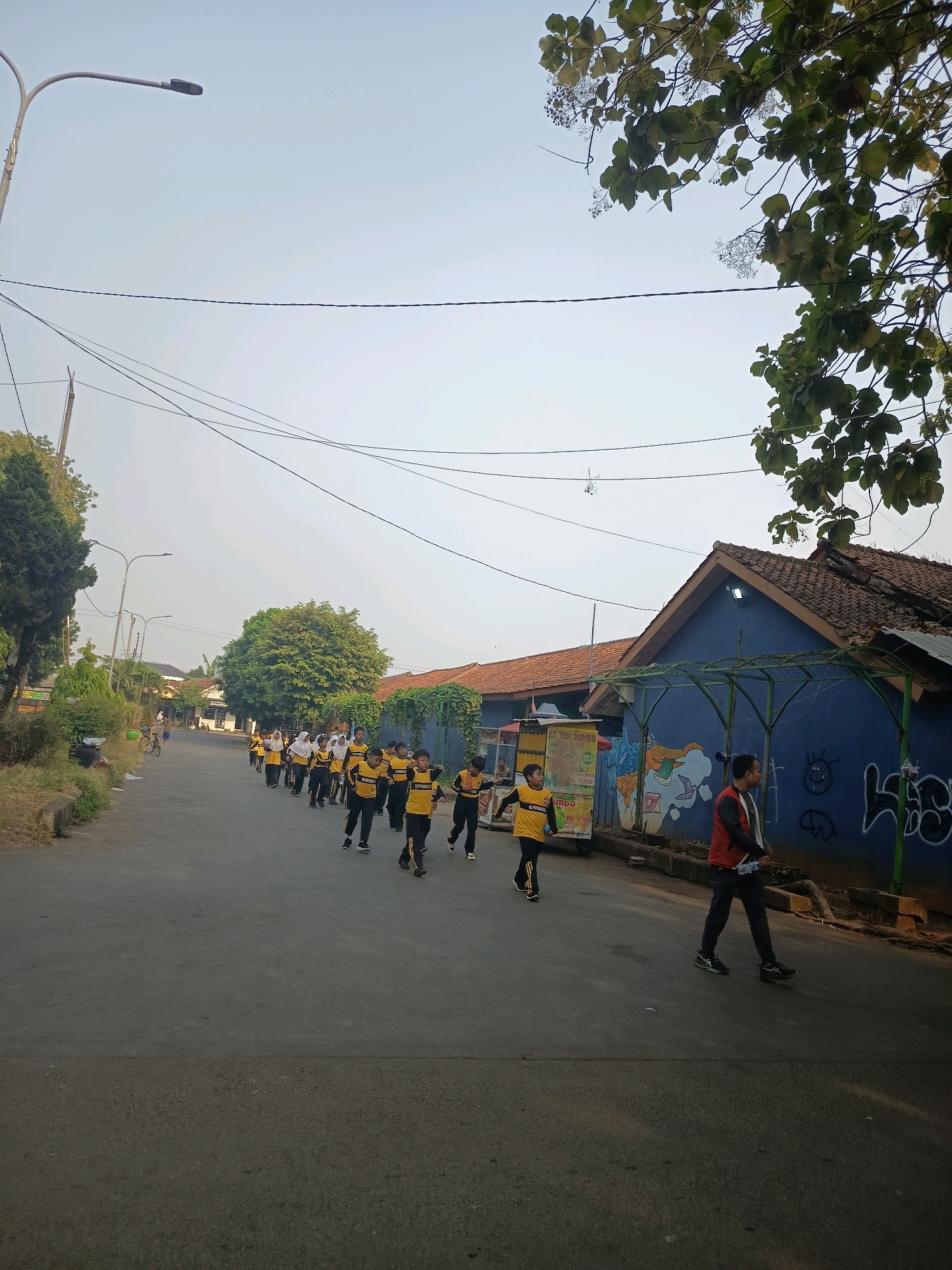 A group of children in yellow and black uniforms are walking in a line down a paved street. They appear to be in motion, possibly participating in a school or athletic event. Some of the children are wearing white headscarves. A few adult figures are also present, including one man in a red vest walking in the foreground on the right side of the street. The street is lined with trees and buildings. One building is painted blue and has graffiti on its wall. There is a food cart to the left of the street. Overhead, power lines stretch across the frame, and a lamppost is visible on the left side of the image. The sky is clear and bright, suggesting it is daytime.