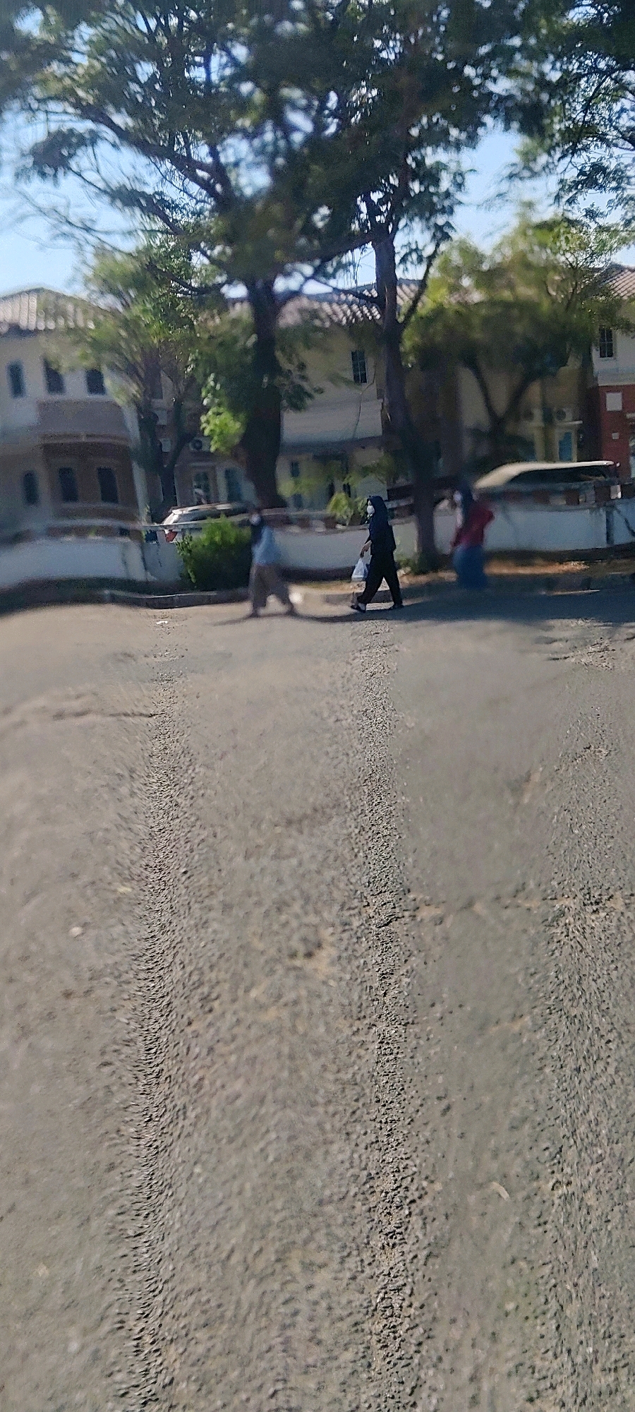 In Mekarmukti, Indonesia, three individuals are depicted walking on a paved road in front of buildings. The person on the left is wearing a light-colored top and pants and is partially obscured by a tree. In the center, another individual, dressed in a dark outfit and a hijab, carries a white bag in their right hand. The person on the right is wearing a red top and dark pants. All three individuals appear to be wearing face masks. The background features multi-story buildings with visible windows and tiled roofs, set against a backdrop of trees and a clear sky. The road in the foreground shows tire tracks, suggesting vehicular activity.