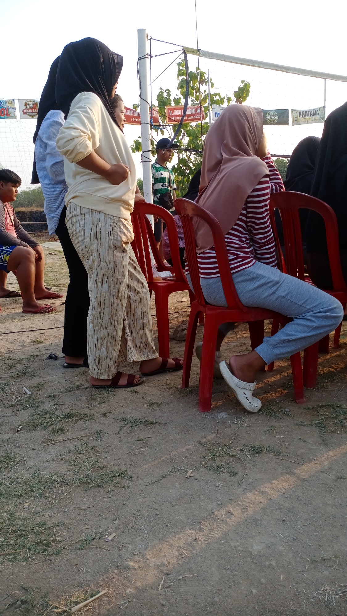 The image shows a group of people in what appears to be an outdoor gathering or event, possibly in Mertan, Indonesia. In the foreground, several individuals are visible. A person wearing a black hijab and a light-colored long-sleeved shirt and patterned pants is standing, their left arm bent. To their left, another person in a light blue and white striped shirt is partially visible. To the right, a person wearing a light pink hijab and a striped red and white long-sleeved shirt is seated on a red plastic chair, wearing grey pants and white clogs. Another red plastic chair is next to them. Further in the background, there are banners with text and images, and a boy in a green and white striped shirt is standing. The ground is dirt with some dry grass. The overall lighting suggests it is daytime, possibly late afternoon, with sunlight casting shadows.