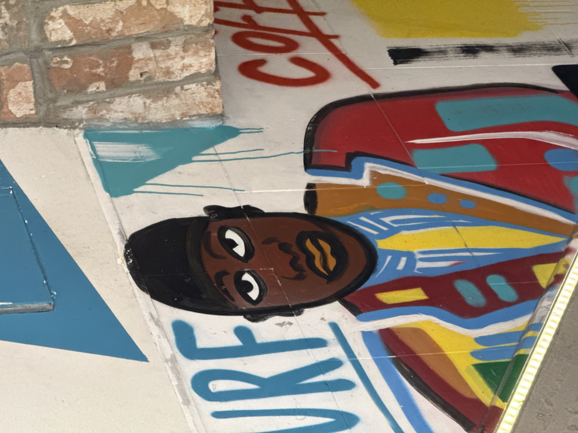 The image shows a section of a graffiti mural on a wall, possibly an underpass or tunnel. The mural features a stylized depiction of a person's face with dark skin, wearing a black hat, and adorned with abstract patterns and colors in red, blue, yellow, and brown. The word "SURF" is written in large blue letters near the face. The word "COFFEE" is written in red above the character. Part of a brick wall is visible in the upper left corner. There is a blue triangle near the left corner. The location is likely Moscow, Russia.