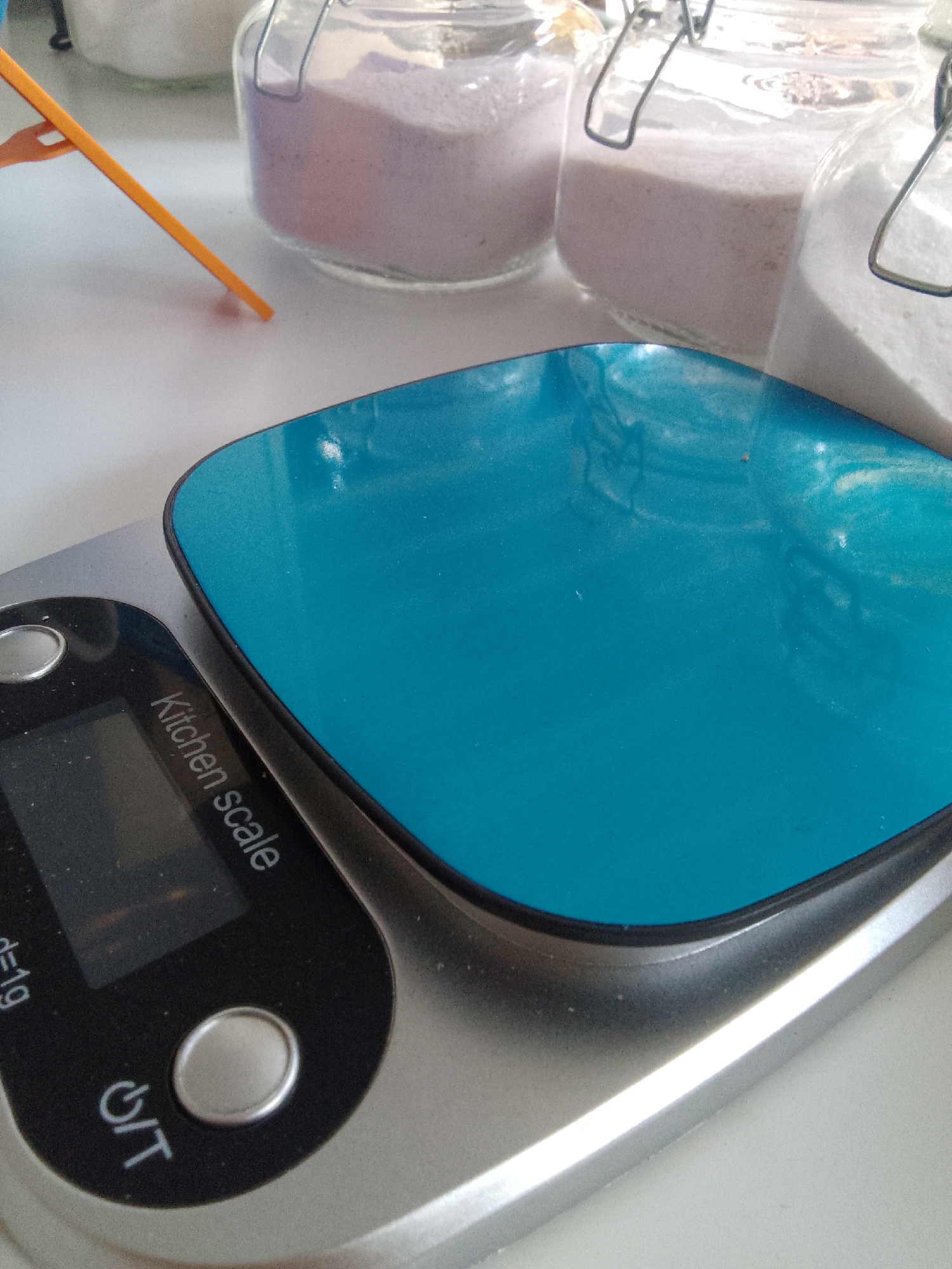 A kitchen scale with a blue weighing surface sits on a white surface. Three glass jars containing powders are behind the scale. An orange utensil rests on the white surface to the left of the scale. The scale has a digital display and buttons. The scene appears to be indoors, possibly a kitchen. There are no people visible in the image. The image does not provide any information about the city of Trenggalek, Indonesia.