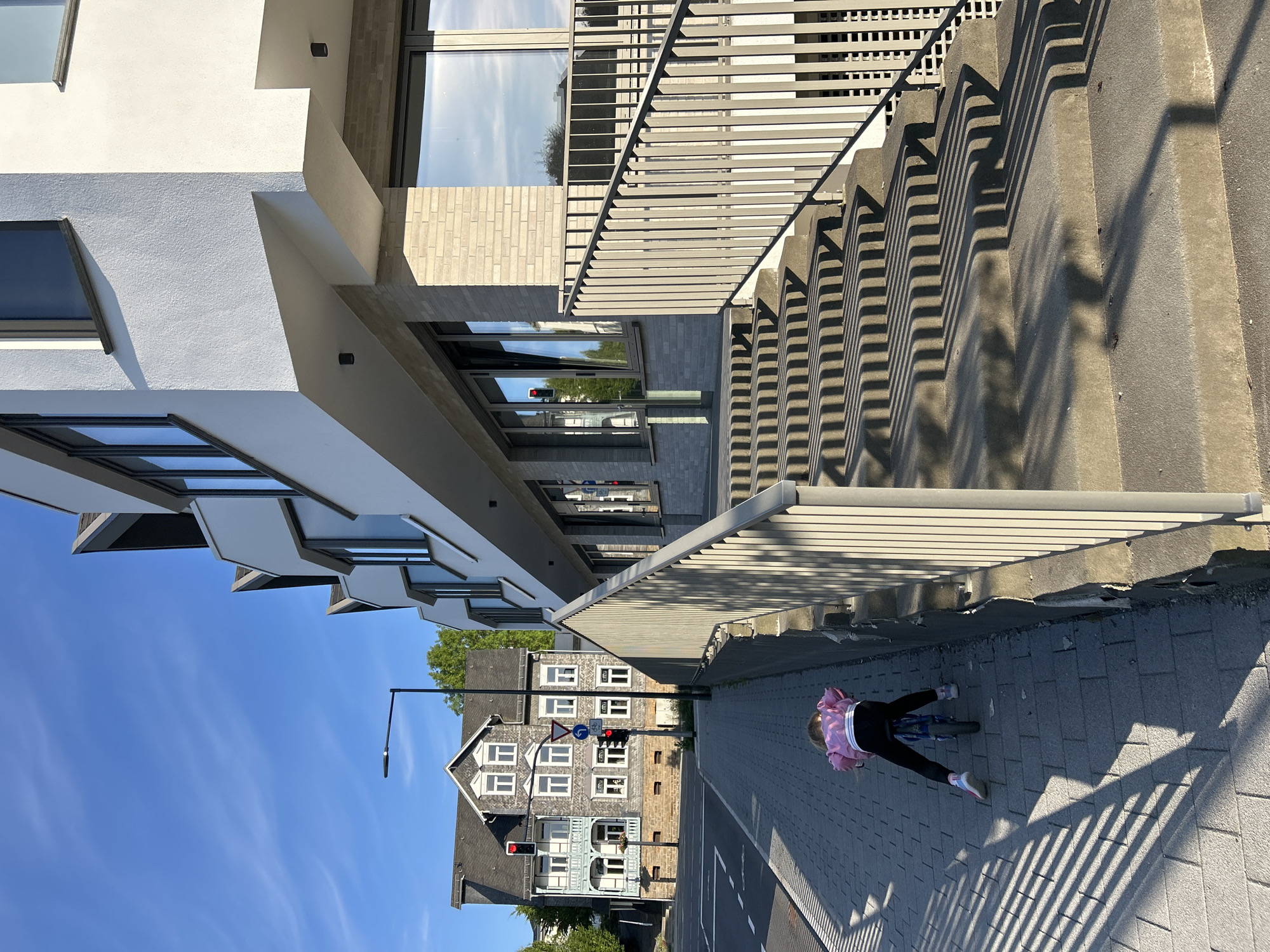 In Waldbröl, Germany, a young child wearing a pink jacket and black leggings is riding a blue bicycle on a grey paved sidewalk. The child is facing away from the camera and is positioned in the lower-left quadrant of the image. To the right of the sidewalk, a set of concrete stairs with a metal railing leads up to a modern, multi-story building with white and grey facades and large windows. The building casts long, striped shadows onto the stairs. In the background, to the left of the modern building, is a traditional German house with a dark roof and multiple windows. The sky is clear and blue with a few wispy clouds, and the sun is shining, casting distinct shadows. A street light is visible to the left of the traditional house. Traffic lights and street signs are present at an intersection in the background.