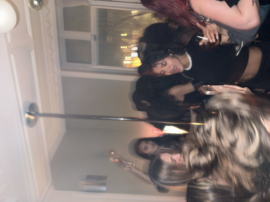 The image depicts an indoor night scene of a crowded social gathering, likely a party, within a building in Paris, France. The room features light-colored walls, ornate crown molding on the ceiling, and a large multi-pane window in the background revealing blurred city lights at night. A prominent, reflective metallic pole extends vertically from the ceiling, positioned centrally within the frame.
Multiple individuals are present, predominantly young women, in close proximity. In the mid-ground, a woman with dark, curly hair, wearing a black crop top that exposes her midriff and a white-collared undershirt, holds a clear glass in her left hand. Her mouth is open, indicative of singing, shouting, or laughing loudly. To her left, partially obscured, is another woman with light brown hair, smiling. Further left in the background, a woman with reddish-brown hair is visible, raising a clear champagne flute in her right hand.
In the foreground, blurred hair from additional individuals indicates their closeness to the camera, with one person holding a light-colored smartphone pointed towards the central group. To the right of the central figure, a woman with reddish hair is partially visible, carrying a dark shoulder bag. The scene is dimly lit by ambient light and reflections, including some colored light sources in the background, contributing to a lively and energetic atmosphere.
