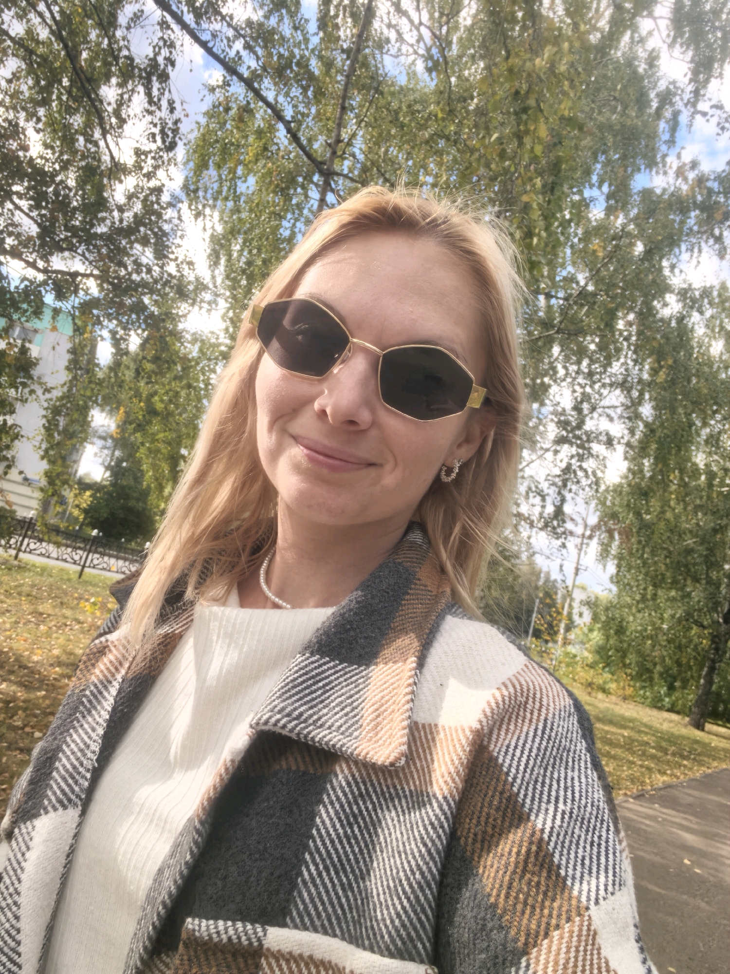 In Sterlitamak, Russia, a woman with blonde hair and sunglasses stands outdoors. She is wearing a white top and a plaid coat in shades of gray, white, and brown. A pearl necklace is visible around her neck. The background consists of trees with green and yellowing leaves, indicating an autumn setting. Fallen leaves are scattered on the ground. A sidewalk is visible to the right, and a building and a fence are present in the distance to the left. The sky is partly cloudy.
