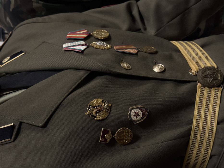 A green military uniform is adorned with various medals and insignia. Ribbons with attached medallions are pinned to the upper chest. Smaller, decorative pins featuring eagles, stars, and Soviet symbols are clustered below. A striped, tan-colored strap with a large, star-shaped button runs vertically along the fabric. Dark shoulder boards are visible, featuring gold accents.