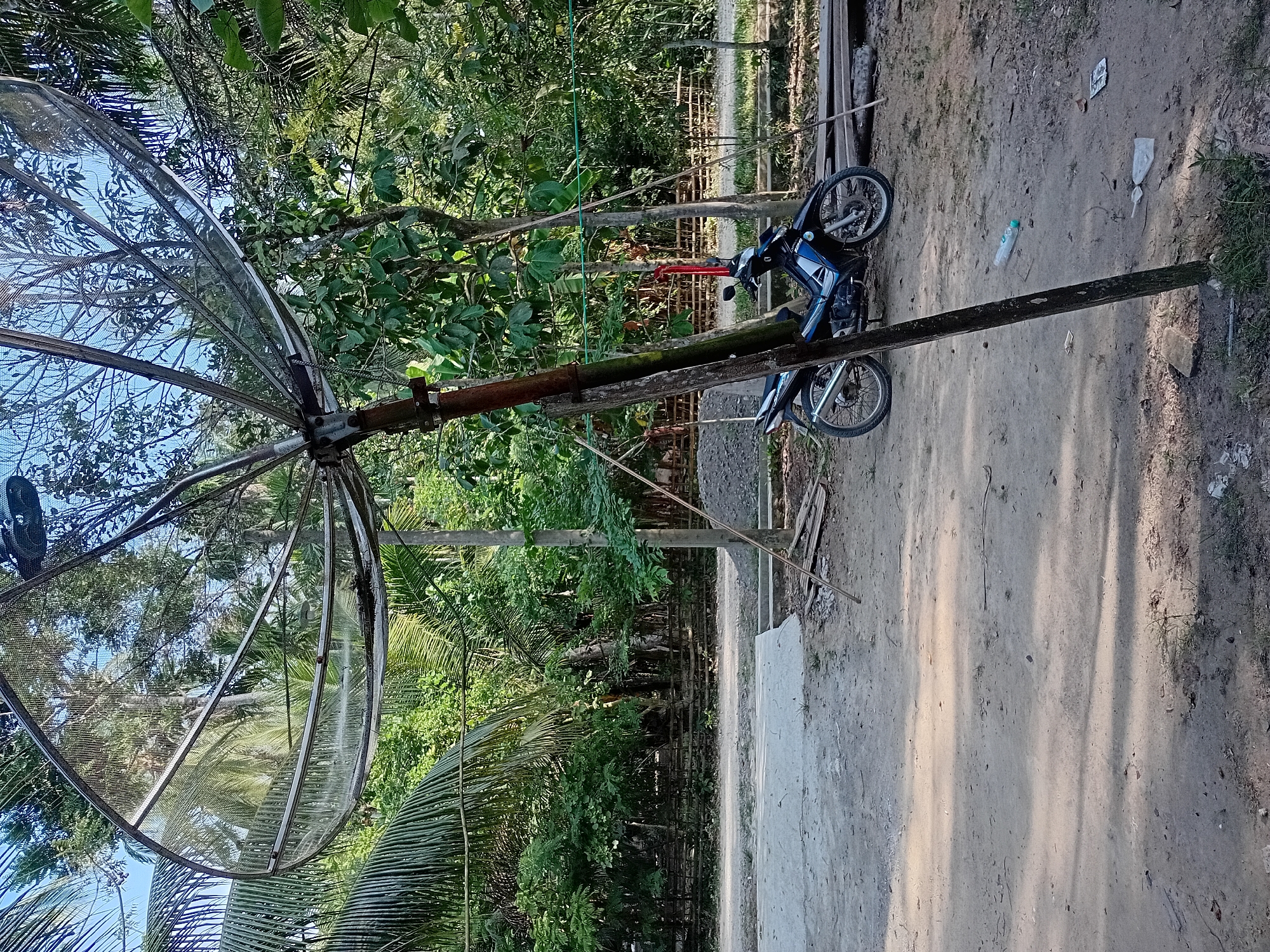In Aluebuya, Indonesia, a satellite dish is erected on a wooden pole, its metallic surface reflecting the lush greenery surrounding it. The dish is positioned at the edge of a dirt path, adjacent to a paved area. A blue and black motorcycle is parked nearby, its tires resting on the uneven ground. In the background, a dense forest of palm trees and other vegetation forms a natural barrier, suggesting a rural or semi-rural setting. Scattered on the ground are a few plastic bottles and wrappers, indicating the presence of human activity. The scene is illuminated by natural sunlight, casting shadows across the path and the objects within the frame.