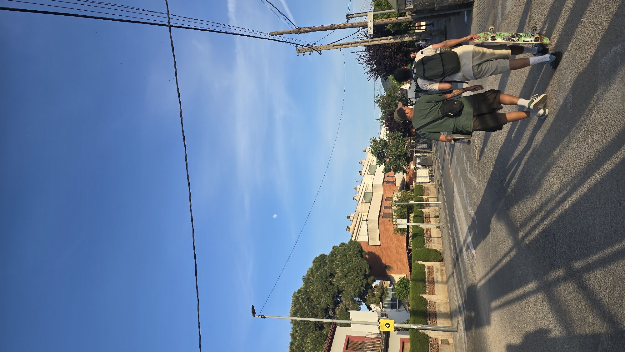 The image depicts a street scene in Santa Perpètua de Mogoda, Spain, on a bright, clear day, likely late afternoon given the long shadows and visible daytime moon. The composition is oriented vertically, showing a wide expanse of blue sky, a paved road, and buildings lining one side.
Two young men are positioned on the asphalt road, possibly on a faded crosswalk. The man in the foreground is dressed in a dark green t-shirt, dark shorts, white socks, and white sneakers with green accents, and wears a dark crossbody bag. A tattoo is visible on his left arm. He holds a skateboard. Behind him, another man wears a light-colored t-shirt, light shorts, white socks, and dark sneakers, carrying a dark backpack with a water bottle. He is also holding a skateboard, the underside of which features a green graphic. They appear to be walking or standing, not actively riding their skateboards.
The right side of the road is bordered by multi-story residential buildings with light-colored facades and some red brick details, interspersed with green hedges and trees. Overhead utility poles and numerous power lines traverse the blue sky. A street lamp pole stands on the left side of the road.
The sky is a clear, vibrant blue with a few subtle white clouds. A distinct crescent moon is visible in the upper left portion of the sky. Long, sharp shadows from the people, poles, and unseen objects stretch across the road surface, indicating the sun is low in the sky. Visible text includes the number "32" on a sign attached to a structure in the background and a yellow directional sign with a black arrow pointing right near the street lamp.