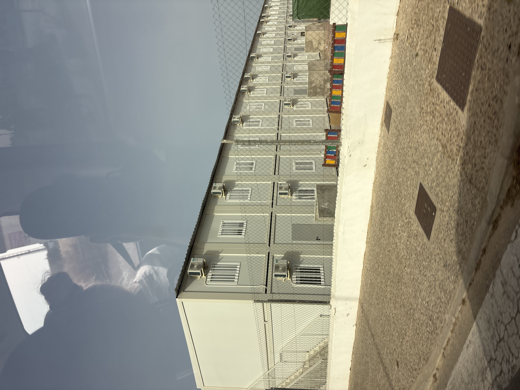 The image shows a row of modular buildings, likely living quarters or offices, constructed from prefabricated panels. These buildings are two stories high and have a uniform appearance with white-framed windows and air conditioning units mounted on the exterior walls. A chain-link fence encloses the buildings, and a white concrete wall runs along the front. In the foreground, a cobblestone or gravel pathway is visible, with several metal grates embedded in it. A colorful fence, made of vertical wooden planks painted in red, yellow, green, and blue, is located behind the white wall. The sky above is clear and light blue. Reflections of people and possibly the interior of a vehicle are visible on the glass surfaces, suggesting the photo was taken from inside a vehicle. The location is identified as Orihuela, Spain.