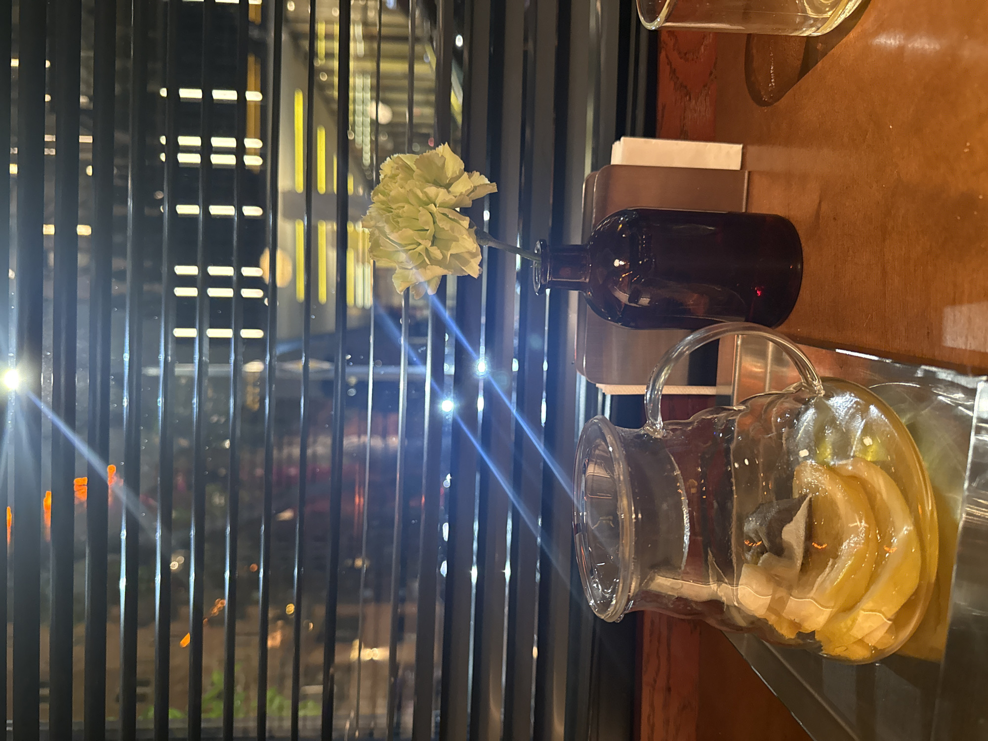 The image captures an indoor still life in Yerevan, Armenia. A clear glass pitcher with slices of lemon and a tea bag is on a dark-brown wooden table beside a small dark-brown glass bottle containing a single light-green flower. Behind the objects is a window covered with black vertical blinds, through which lights from a city nightscape are visible. The scene is softly illuminated, creating a quiet and composed atmosphere.