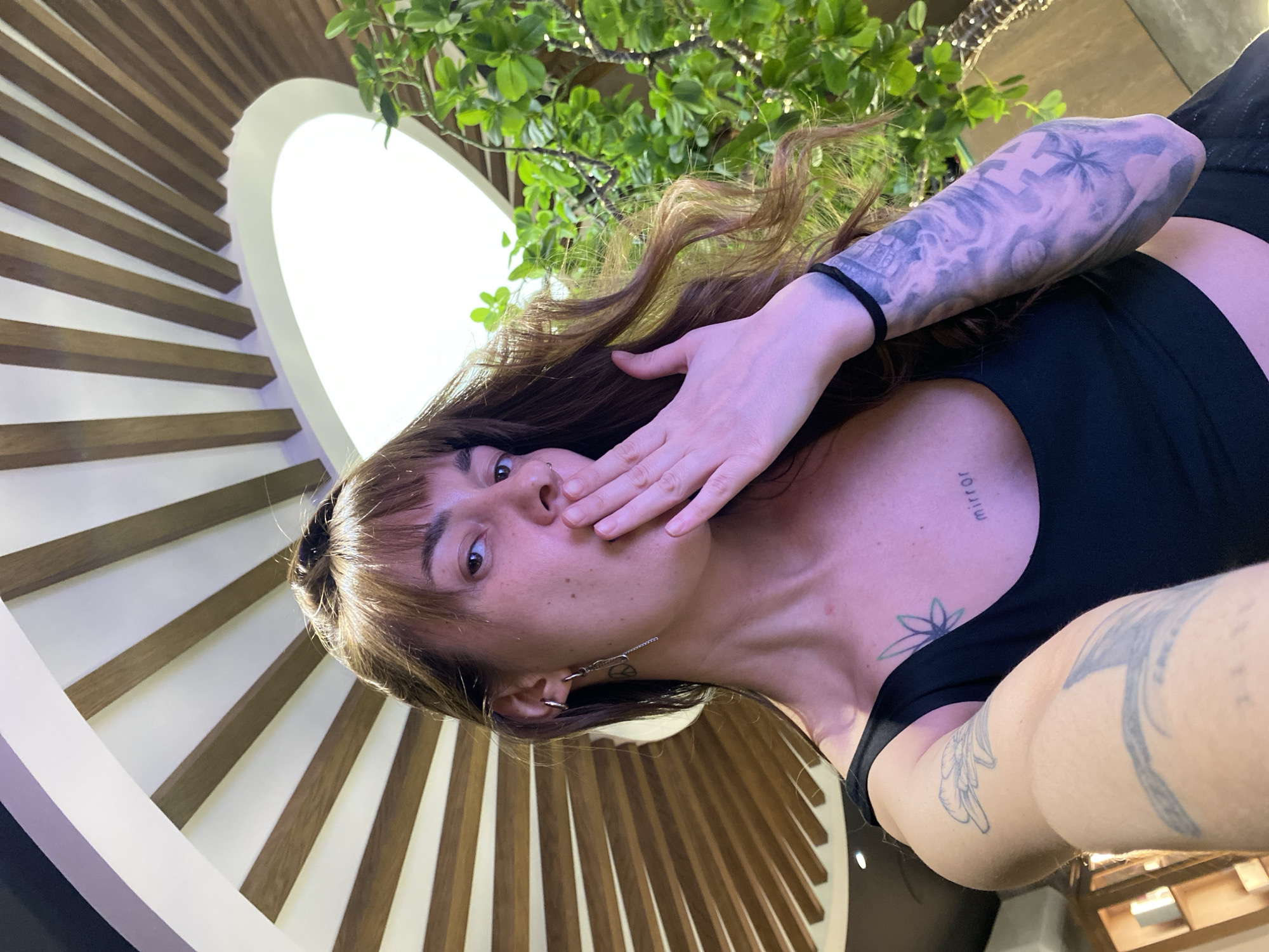 A person with brown hair and bangs is shown in a low-angle selfie, partially covering their mouth with their hand. They are wearing a black top and have tattoos on their arms and chest. The background features a spiraling architectural design with wooden slats and a white border, resembling a modern staircase or a decorative art installation. Green foliage is visible above the person, suggesting an indoor plant or tree element. The location is not identifiable as Votkinsk, Russia based on the visual elements in the image.