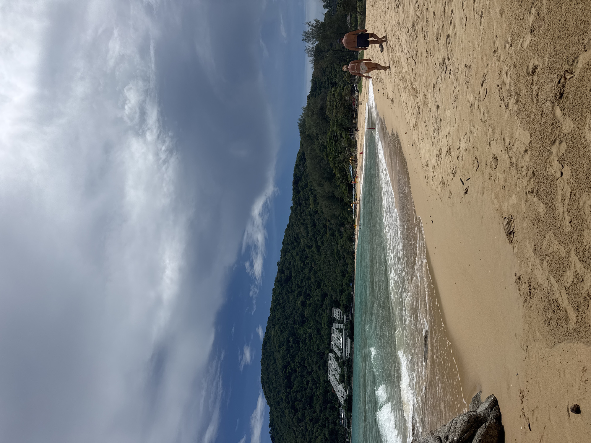 This is a vertical, eye-level, medium shot of a tropical beach with a couple walking away from the camera on the right side of the frame. The couple consists of a woman in a white bikini and a man in dark swim trunks. The beach is composed of light brown sand and the ocean is a vibrant turquoise color with white-capped waves rolling onto the shore. In the background, a lush green, tree-covered mountain rises from the coastline. On the lower slope of the mountain, a series of white buildings are visible, presumably a resort or hotel complex. The sky above is partly cloudy, with patches of bright blue interspersed with dramatic dark gray clouds. The scene conveys a sense of relaxation and natural beauty.