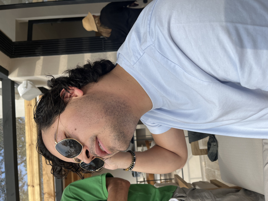 A man with curly black hair and sunglasses tilts his head, facing the camera with a slight smile. He's wearing a light blue t-shirt and a beaded bracelet. The background shows glimpses of an outdoor setting, including a wooden structure, another person in a straw hat, and someone wearing a green shirt. The picture appears to be taken from a slightly off-kilter angle indoors or on a patio.