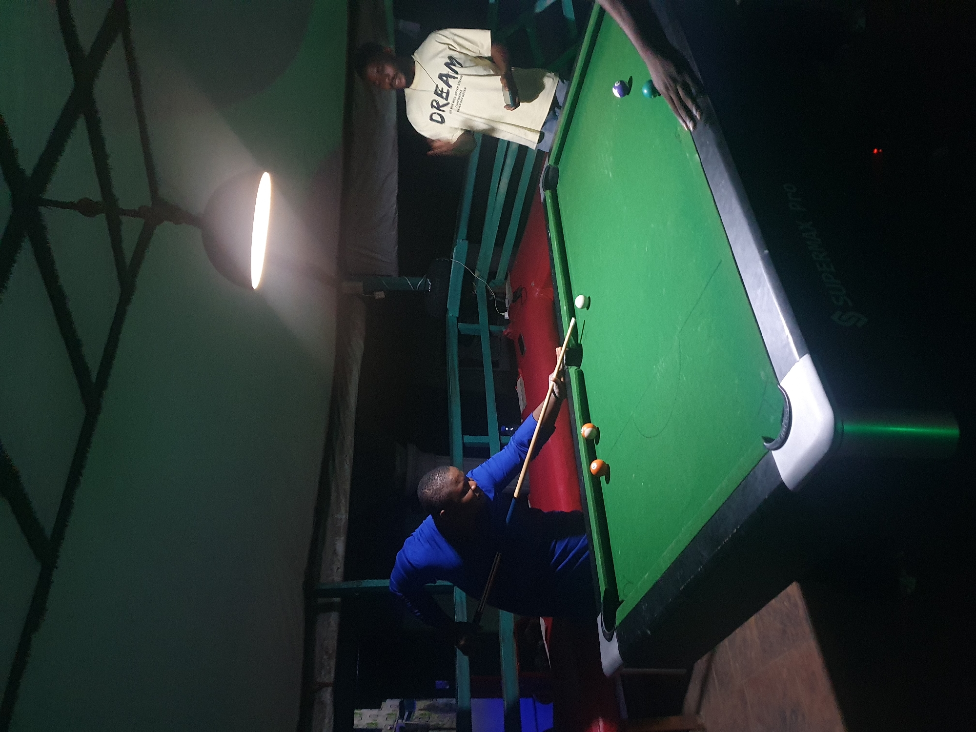 In Lokoja, Nigeria, two men are engaged in a game of pool under a covered outdoor area at night. A pool table with green felt occupies the foreground. One man, wearing a blue long-sleeved shirt, is positioned to strike a cue ball with a pool cue, aiming towards two billiard balls on the table. The other man, in a light-colored t-shirt with the word "DREAM" printed on it, stands to the right, holding a smartphone and gesturing with his other hand, appearing to be observing the game or conversing. A third person's arm is partially visible on the right side of the table, reaching towards some billiard balls. The environment suggests a recreational space, possibly a bar or a private outdoor gathering area, illuminated by a pendant light fixture.