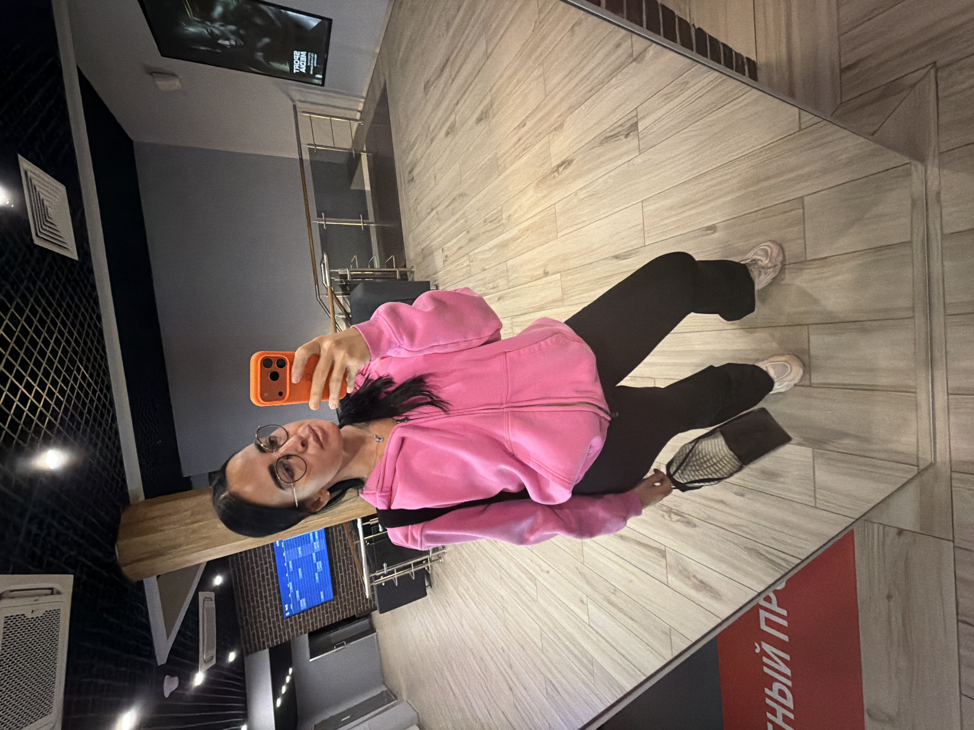 A young woman is captured in a mirror selfie, seen from the waist up, standing on a mirrored floor that reflects her lower body and the surrounding interior. She is wearing a bright pink zip-up hoodie, black pants, and light-colored sneakers. Her hair is dark, and she wears glasses, with a neutral to slightly smiling expression. She holds an orange-cased smartphone in her right hand, positioned to take the picture.
The setting appears to be a modern indoor space, possibly a commercial or public building, consistent with locations found in Saint Petersburg, Russia. The background features light-colored wooden floor tiles that extend onto the mirrored surface, and grey and dark walls. A dark, patterned ceiling with recessed lighting is visible in the reflection.
Notable objects include a metal railing or shelving unit, an air vent on a dark wall, and a large screen mounted on the ceiling, reflecting a partially visible word, likely "WEDIV" (reversed, possibly "VIDEO") and a number "700". To the woman's left, a wooden plank or shelf is visible, and another screen further back displays blue graphics or text against a brick-like texture. A black handbag hangs from her left shoulder. On the mirrored floor, a red and black sign with white Cyrillic text is visible in reflection, showing the reversed fragment "Й ПР", suggesting a word like "ОБРАТНЫЙ ПРО..." (meaning "reverse pro..." or "return pro...").
The activity taking place is a self-portrait taken in a full-length mirror, utilizing the reflective properties of the floor to enhance the visual composition. The time of day is not explicitly indicated but artificial indoor lighting is prevalent.