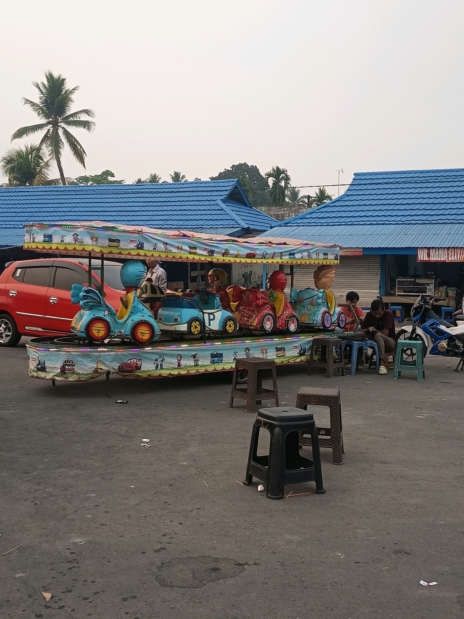 The image shows a small amusement park ride with colorful cars and cartoon characters on a paved lot in Pulau, Indonesia. The ride is under a covered canopy adorned with lights and illustrations. A red car is parked to the left of the ride. In the background, there are buildings with blue roofs, and palm trees are visible in the distance, suggesting a tropical setting. Several people are present. Two individuals are seated at a small table with blue chairs, looking down at their phones or papers. Another person is standing near the ride's control panel. A motorcycle is parked to the right. The ground is asphalt, with scattered litter and a few plastic stools in the foreground. The sky is overcast.
