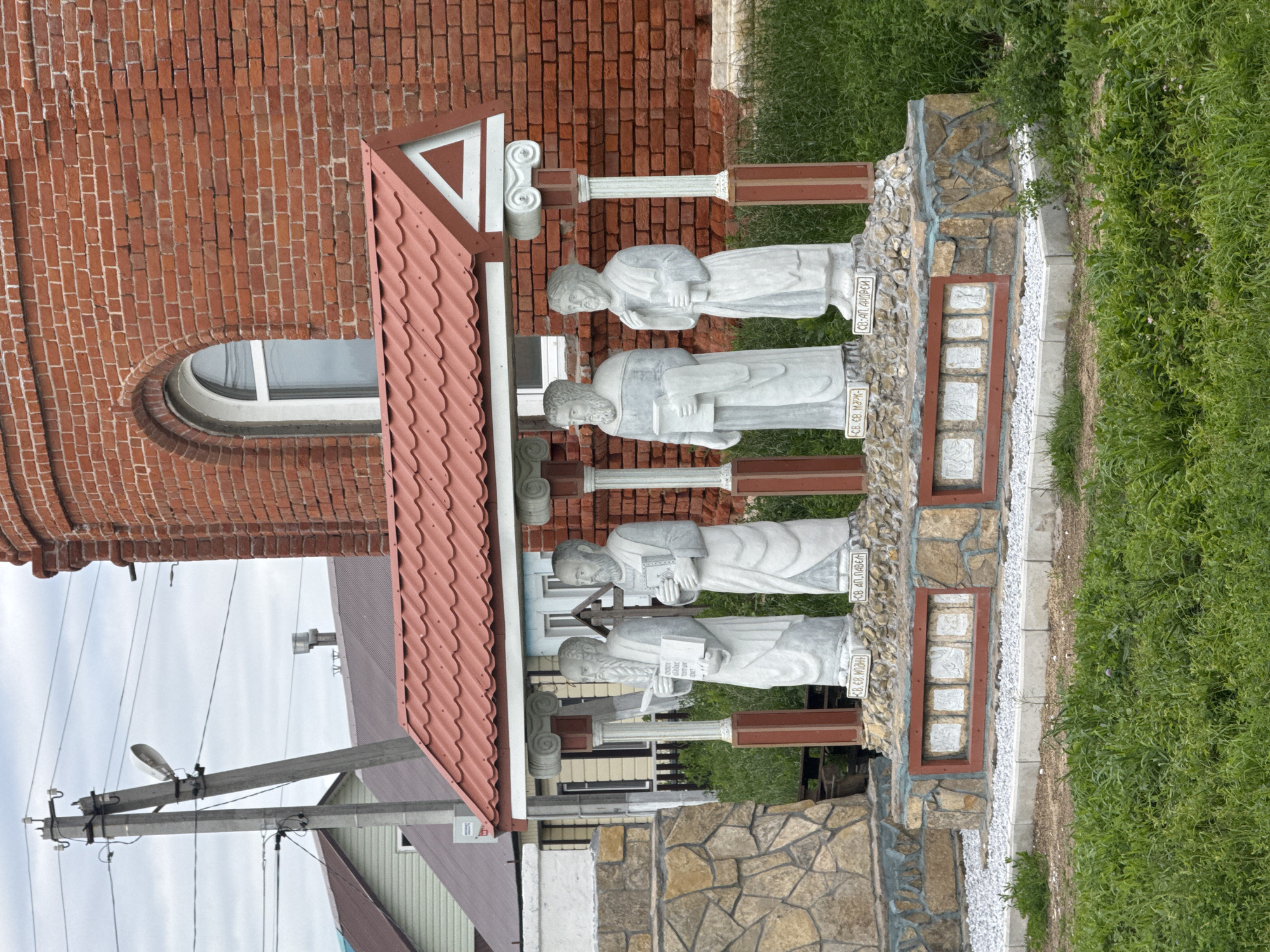 The image shows a religious monument in the city of Anatiš, Russia, featuring four white marble statues of men in togas, each holding a book. The statues are arranged in a row under a red-roofed canopy supported by red and white columns. Each statue has a nameplate at its base. The monument is situated outdoors, with a brick building to the right and a stone wall on the left. Green grass and some gravel surround the monument. In the background, there are houses and utility poles. The sky is overcast.