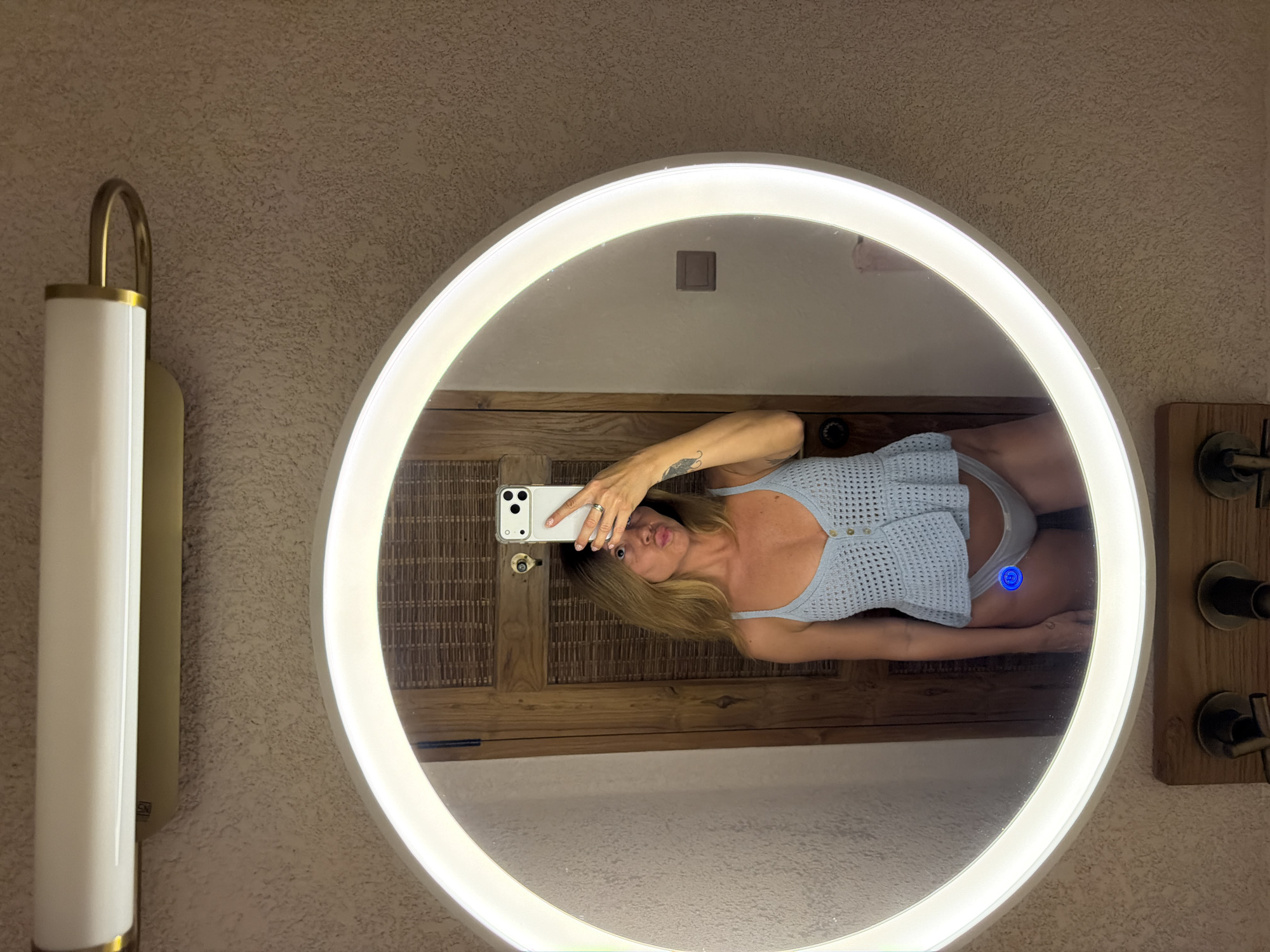 A woman is captured in a mirror selfie within a bathroom or vanity area in Mengwi, Indonesia. The image is rotated 90 degrees clockwise, showing the woman lying on her side in the reflection. She has long blonde hair and is wearing a light blue, crocheted or knitted sleeveless top and light-colored underwear or swimwear bottoms. She holds a white iPhone with her right hand to take the photo, and her left arm is visible, showing a small tattoo resembling a feather or wing design on her forearm. She makes a playful "duck face" expression towards the camera.
The mirror is circular and features an integrated bright LED light strip around its perimeter, which illuminates the subject. A small, illuminated blue circular button is visible on the mirror's surface in the reflection, likely an activation switch for the LED light or a defogger.
The background visible around the mirror and in its reflection suggests an indoor setting. The wall has a textured, light-colored finish, possibly stucco. To the left of the mirror, a modern wall sconce with a long, cylindrical white shade and brass accents is mounted. The reflection behind the woman shows a rustic wooden surface, possibly a slatted cabinet or door. On the right side of the mirror, outside the frame of the reflection, elements resembling dark-colored faucet handles on a wooden panel are partially visible. The overall lighting suggests an indoor, possibly evening, setting with the mirror's light being a primary source of illumination. No discernable text is visible.