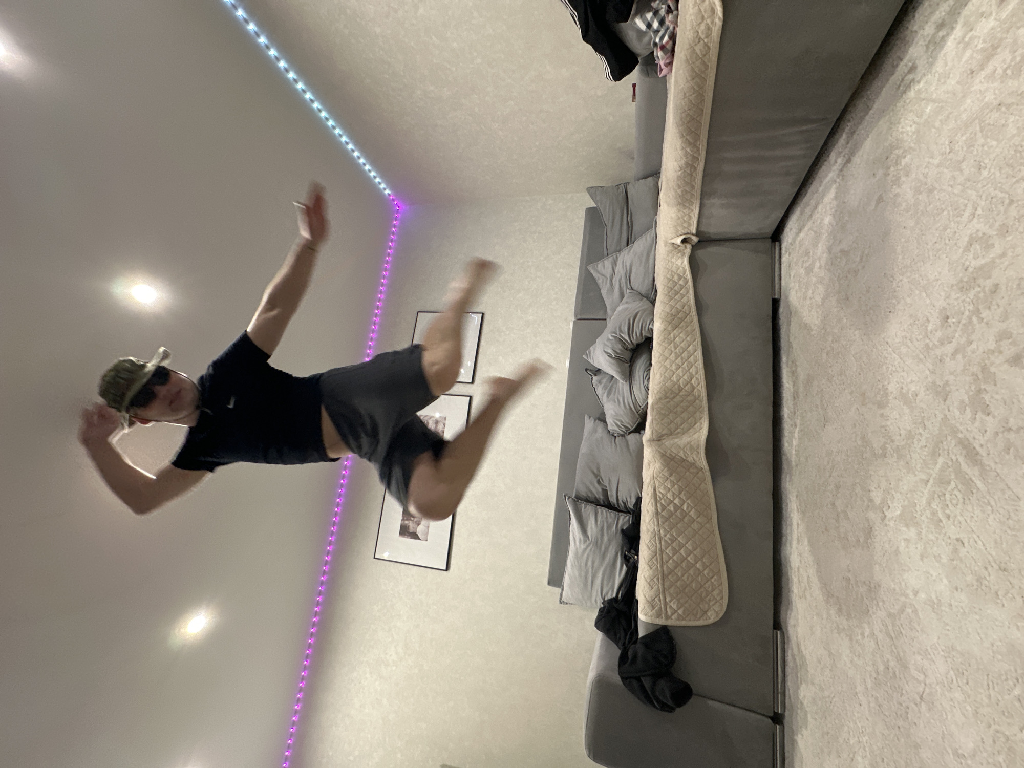 The image displays an indoor scene, presented sideways, with the room's floor along the right edge and the ceiling along the left. A young person is captured in mid-air, suspended horizontally across the frame, suggesting a jump, fall, or acrobatic move.
The main subject is a male, likely a young adult or teenager, wearing a dark t-shirt, dark shorts, sunglasses, and a camouflage-patterned cap. He has one arm raised and his legs bent, indicating motion, with a slight blur to his body.
The setting is a modern living room or lounge area. On the right side of the image, a large, grey sectional sofa with multiple cushions is visible. A light-colored, quilted throw blanket is draped over a section of the sofa. The actual floor, positioned along the right edge of the frame, is covered with a light-colored, textured rug or carpet. The wall directly behind the person (which appears as the 'ceiling' in this orientation) features two framed pictures with black frames and white matting, though their content is indistinguishable. Running along the corner where the wall meets the ceiling, there are two strips of LED lights, one emitting a blue/white glow and the other a purple glow, providing accent lighting. Two other spotlights are also visible on the ceiling. The walls are light-colored and appear to have a textured finish.
The dynamic pose of the person against the contemporary interior suggests a playful and energetic moment, likely photographed in the evening or at night given the prominent use of artificial lighting. No visible text is present in the image.