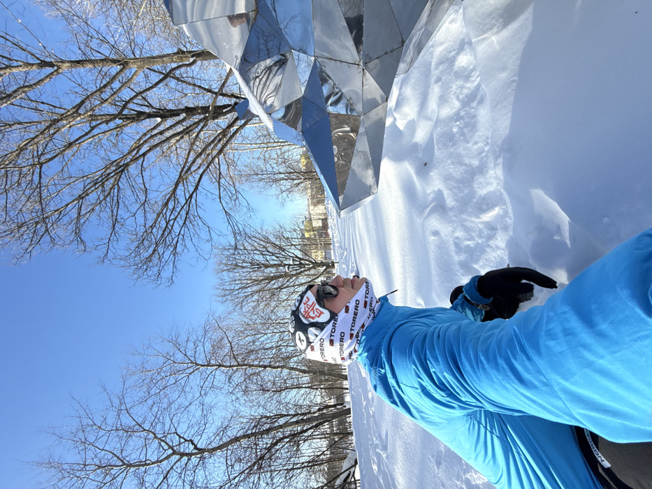 The image captures a winter scene in Millepark (Millennium Park) in Kazan, Russia, under a clear blue sky. A single individual is positioned in the left foreground, appearing to crouch or sit in deep, uneven snow. This person wears a bright blue padded winter jacket, black gloves with blue accents on the cuffs, dark sunglasses, and a white neck gaiter or balaclava patterned with "TORERO" text and "АК БАРС" (AK BARS) in Cyrillic on the forehead area. The individual is looking intently towards the right, specifically at a large, geometrically faceted metallic sculpture, which resembles the head of a snow leopard (Ak Bars). This reflective sculpture is situated in the mid-ground, partially obscured by accumulated snow. The background features numerous deciduous trees with bare branches, indicative of winter, and distant buildings are faintly visible between the trees. The scene is brightly lit by direct sunlight, casting reflections on the metallic sculpture and illuminating the snow.