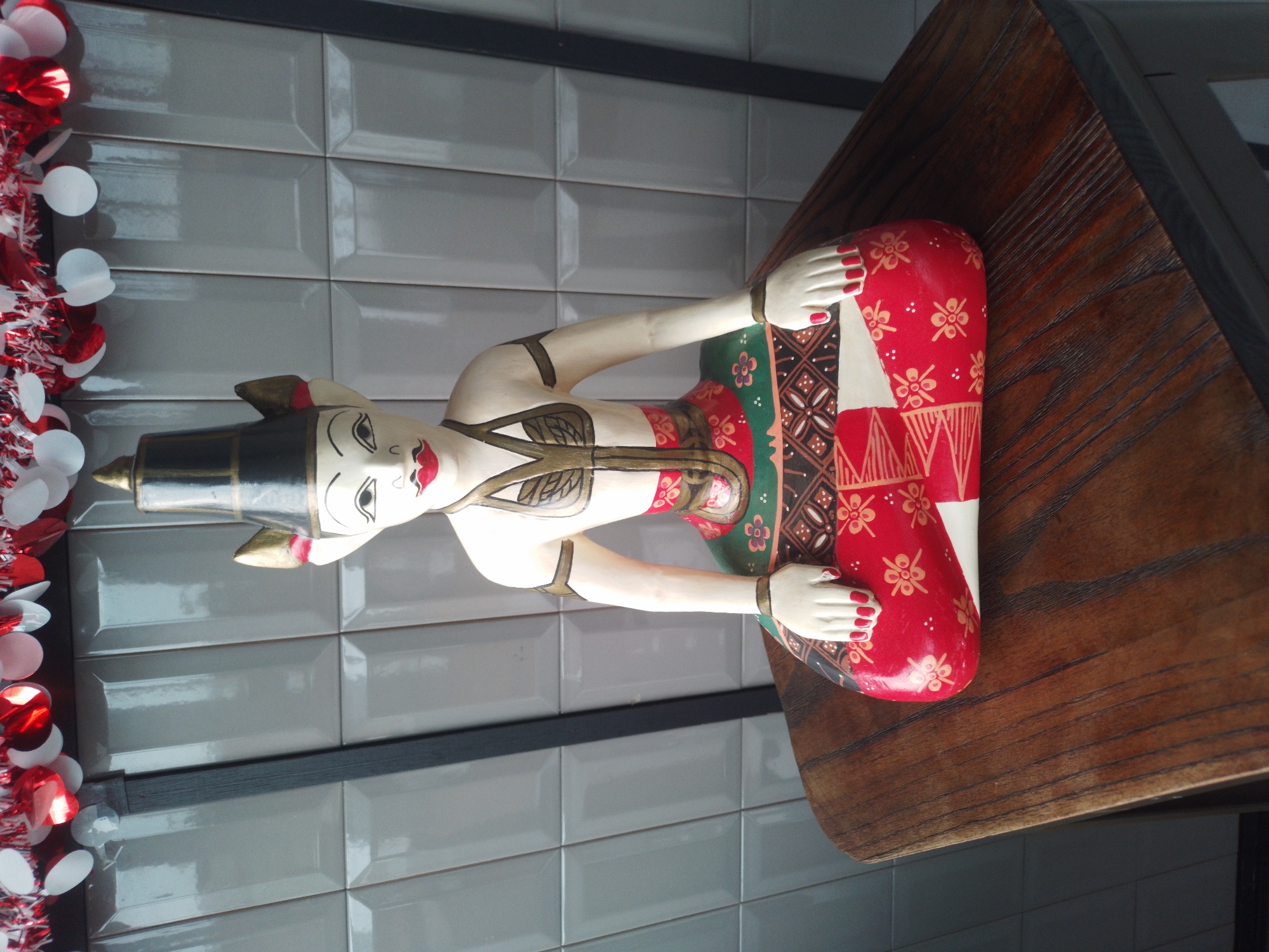 A full-shot, eye-level, medium-light outdoor shot depicts a wooden figurine of a person, possibly a puppet or statue, sitting cross-legged on a wooden table. The figurine is dressed in traditional attire, with a pointed black hat adorned with gold trim and red accents. The body is painted white with intricate gold and red patterns on the chest and abdomen, and the lower body is clad in a colorful sarong-like garment featuring red, green, and black geometric and floral designs. The figurine's hands are resting on its knees, with painted red fingernails.
The background consists of light gray tiled walls, with vertical dark gray supports visible on the left and right. At the top of the frame, a garland of red and white tinsel and circular paper decorations is strung across. The wooden table surface is richly textured, with a dark brown, varnished finish showing prominent wood grain. The scene appears to be set in an open-air environment, possibly a patio or cafe area, in the city of Bumirejo, Indonesia.