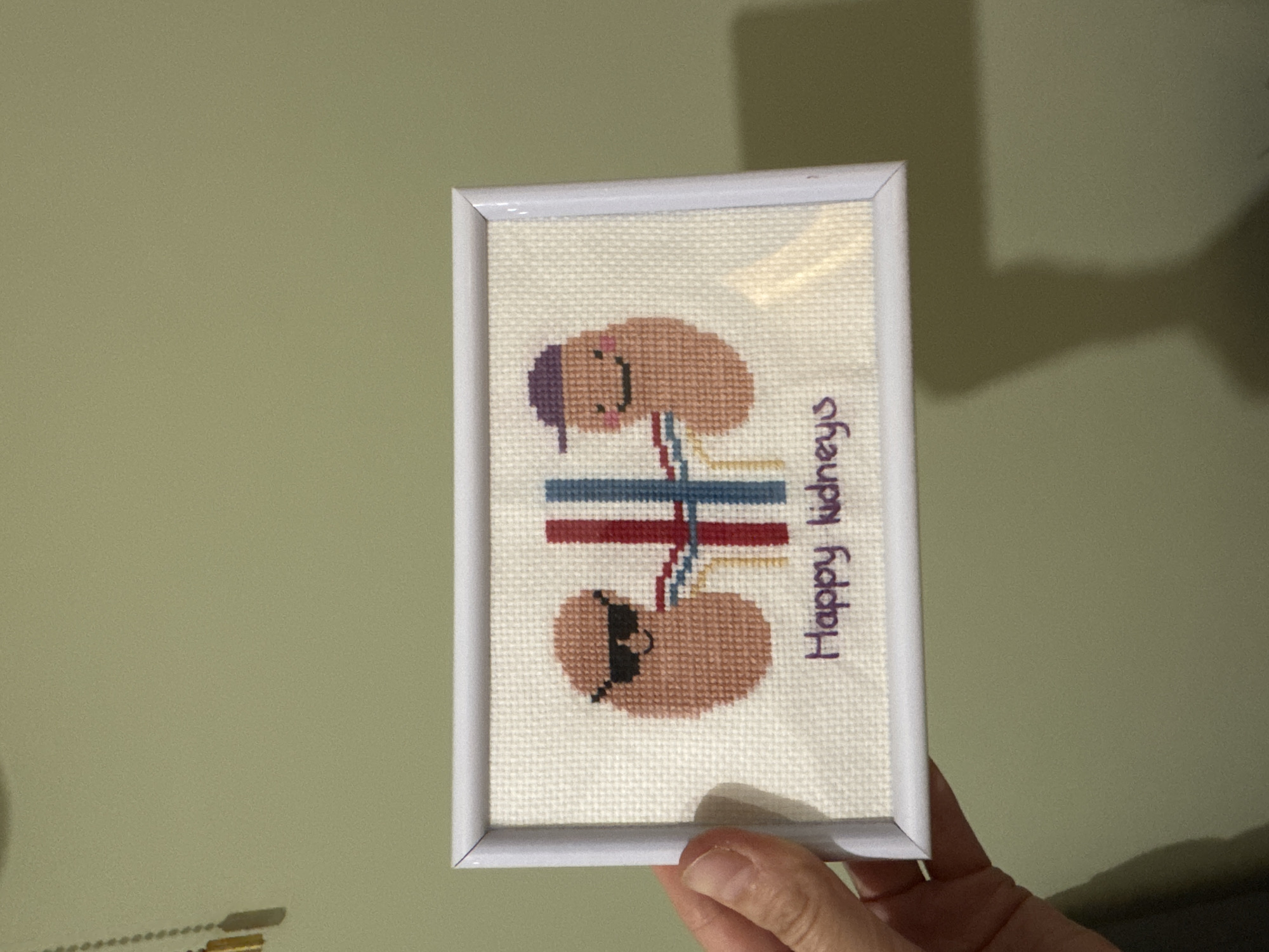 A framed cross-stitch artwork featuring two cartoon kidneys is held by a person's hand against a pale green wall. The top kidney wears a purple hat and smiles. The bottom kidney wears sunglasses. Between them are colored stripes and the phrase "Happy Kidneys" stitched in purple. The frame is white. The location is unspecified, and there is no indication it is in Izhevsk, Russia.