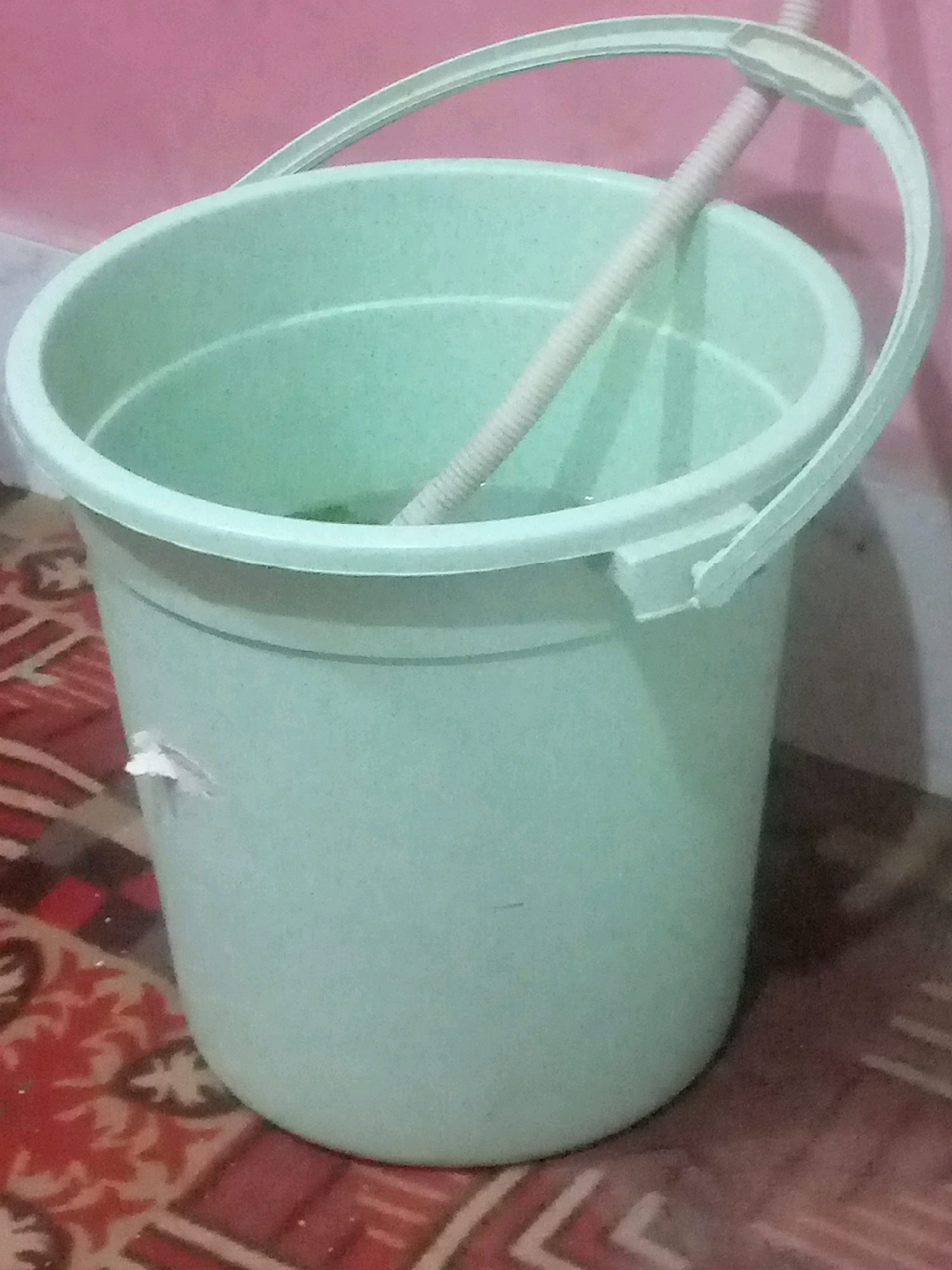 A light green plastic bucket, approximately half-full of water, sits on a patterned rug. A light pink, ribbed hose runs diagonally across the top of the bucket, partially submerged in the water. The bucket has a white plastic handle. A small, white piece of tape is affixed to the exterior of the bucket, near the base. The background consists of a pink wall. The scene is indoors, likely in a domestic setting in Khairpur, Pakistan.