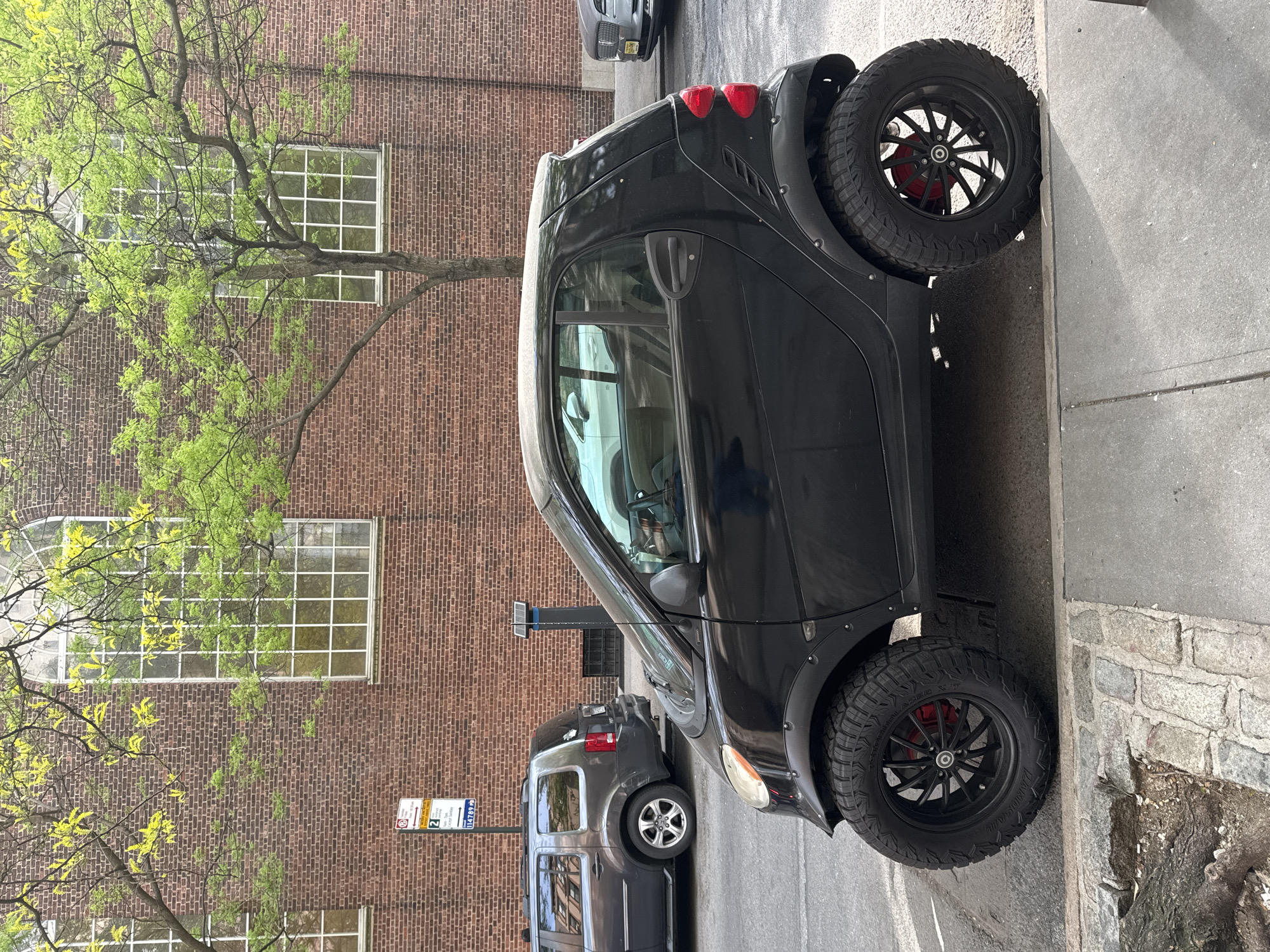 The media file displays a black, heavily modified Smart Fortwo car parked on a city street. The car is equipped with large, aggressive off-road tires on black multi-spoke rims, featuring red brake calipers and extended fender flares. A grey SUV, possibly a Honda Element, is parked directly behind the Smart car, with another grey vehicle partially visible further down the street.
The background consists of a multi-story brick building with several large windows, some exhibiting a grid pattern. A tree with vibrant green, freshly sprouted leaves partially frames the building in the foreground. The scene is illuminated by natural daylight, suggesting it is daytime with clear or partly cloudy weather.
The setting is an urban environment, likely in New York City, characterized by asphalt paving, a stone sidewalk, and street infrastructure. A metal pole on the sidewalk holds multiple white and blue street signs. Visible text on these signs includes "NO STANDING 8AM-7PM MON-FRI", "2 HOUR PARKING 8AM-7PM MON-FRI", and a code "114769 M". A red "P" with a line through it also indicates parking restrictions. The word "LANDSAIL" is discernible on the sidewall of the Smart car's tires. The image itself is presented in a portrait orientation, appearing rotated clockwise from a standard landscape view.