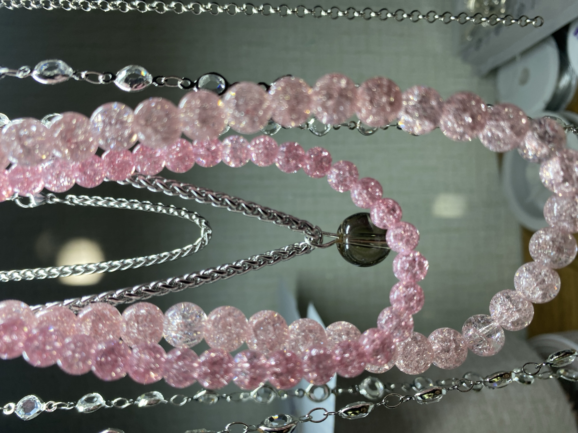 The image shows a close-up of several layered necklaces or bracelets. The primary focus is on a strand of pink, translucent, faceted beads that appear to be made of glass or a similar material. These pink beads are interspersed with smaller, clear, faceted beads and appear to be strung together on a fine thread.
Behind the pink beads, there are several silver-colored chains. One chain is a thicker, more ornate, braided-style chain, while another appears to be a more standard link chain. Hanging from one of the silver chains is a single, dark, translucent round bead, possibly smoky quartz or glass.
To the left of the pink beaded strands, another strand of alternating clear, faceted beads and small silver-colored rings is visible. Further to the right, more silver chains are partially visible.
The background is softly blurred and appears to be a light-colored, possibly reflective surface, with some indistinct shapes and light sources. There are also some spools of thread or wire visible in the lower right portion of the image, suggesting a crafting or jewelry-making context. The overall lighting is soft, highlighting the sparkle and translucence of the beads. There are no people or animals present in the image. The scene is an indoor setting, likely a display or workspace. The location of Kushchovskaya, Russia is not discernible from the visual information provided in the image.