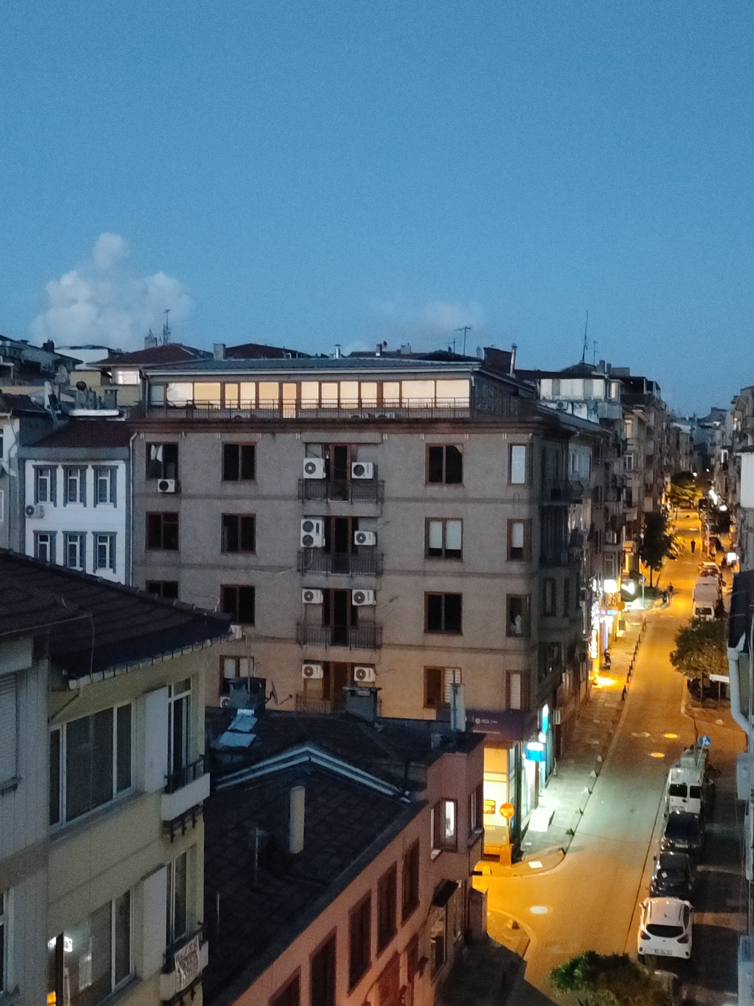 The image captures an urban street scene in Kadıköy, Turkey, during twilight. The perspective is from an elevated position, looking down onto a narrow street lined with multi-story buildings. The buildings exhibit a mix of architectural styles, with some featuring balconies adorned with air conditioning units. The street is illuminated by warm, artificial lighting from streetlights and shop windows, creating a contrast with the deep blue sky. Several vehicles, including cars and a van, are parked along the street. A few pedestrians are visible walking along the sidewalk. The overall atmosphere is one of a bustling city transitioning into the evening.
