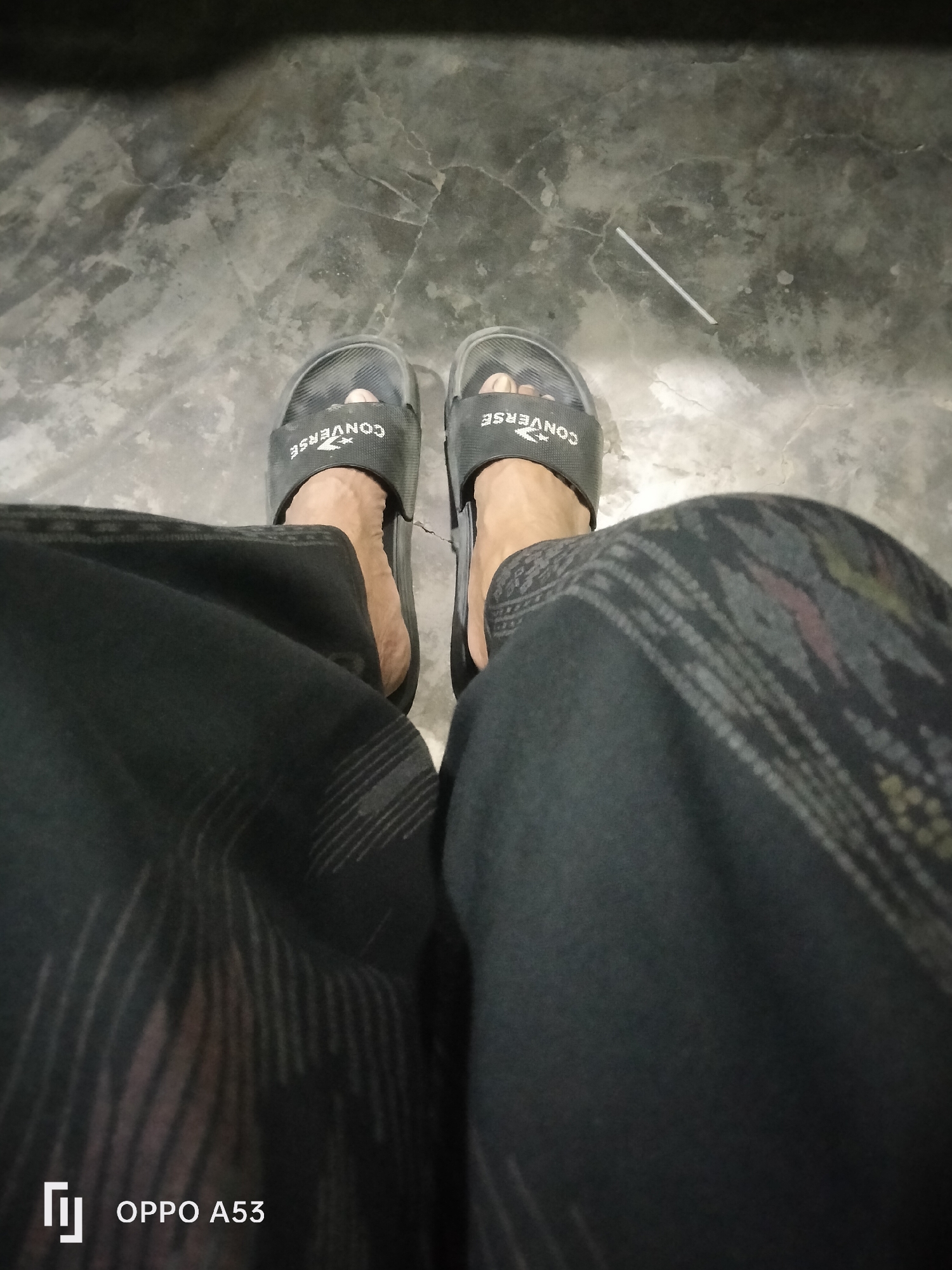 A person is sitting on a floor, their legs are visible from the knees down. They are wearing black Converse slides and dark pants with a patterned design. The floor appears to be concrete or stone with visible cracks and stains. A white object, possibly a piece of straw or plastic, is on the floor. The bottom left corner of the image shows an "OPPO A53" watermark, indicating the photo was taken with this device. The scene is likely indoors, possibly in a home or public space in Badung, Indonesia.
