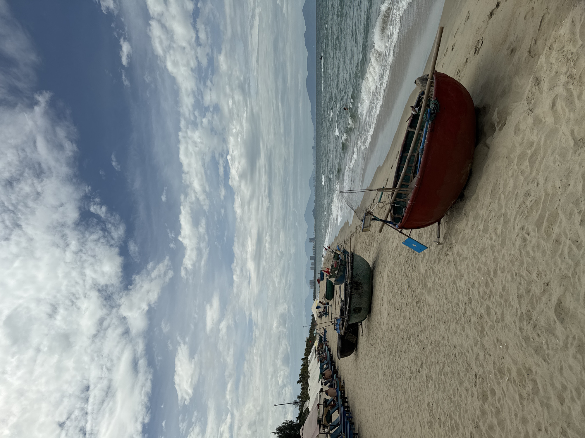The image depicts a serene beach scene in Hoi An, Vietnam, with a focus on traditional Vietnamese basket boats resting on the sandy shore. In the foreground, a vibrant red basket boat is positioned at an angle, its weathered surface contrasting with the light-colored sand. A blue flag and various ropes are visible within and around the boat. Further down the beach, several other basket boats, one painted green, are lined up. Beach umbrellas and lounge chairs are visible on the left, with a few people relaxing. The ocean stretches out to the horizon, with gentle waves lapping the shore. Distant mountains and a cityscape are faintly visible across the water, under a sky filled with dynamic, white clouds against a clear blue backdrop. The overall atmosphere is peaceful and captures a typical coastal day in Hoi An.
