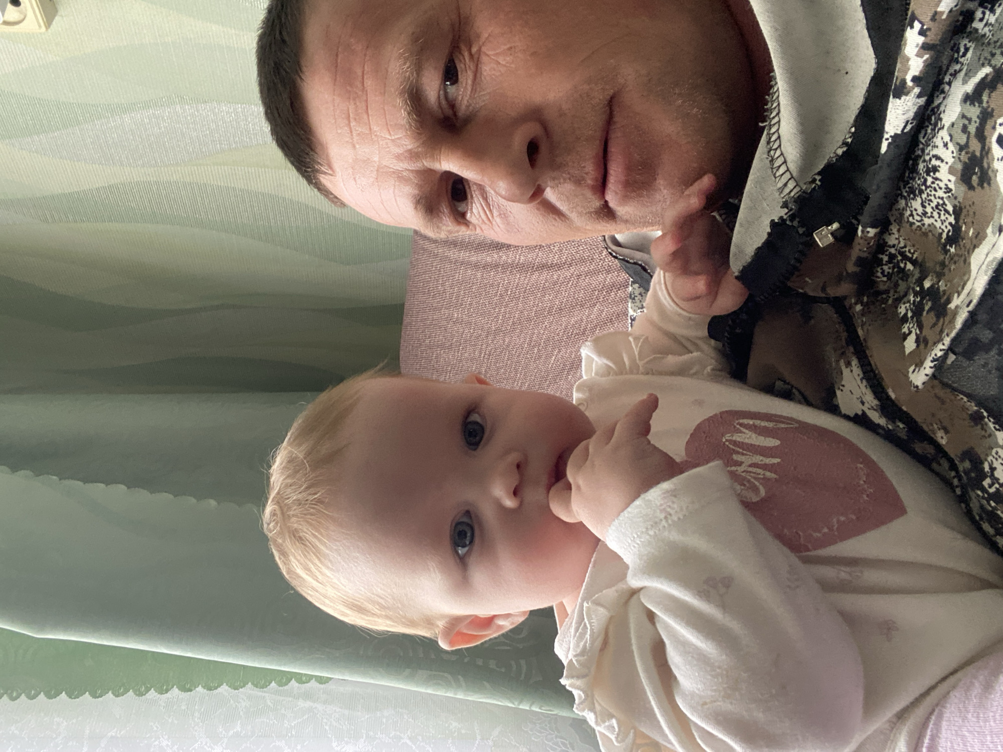 A man and a baby are depicted indoors. The man, who is likely a father or caregiver, has short hair and is wearing a patterned hooded jacket. The baby has fair hair and is wearing a white outfit with a heart design. The scene is indoors, with a patterned wall visible in the background. The location is Фоки, Russia.