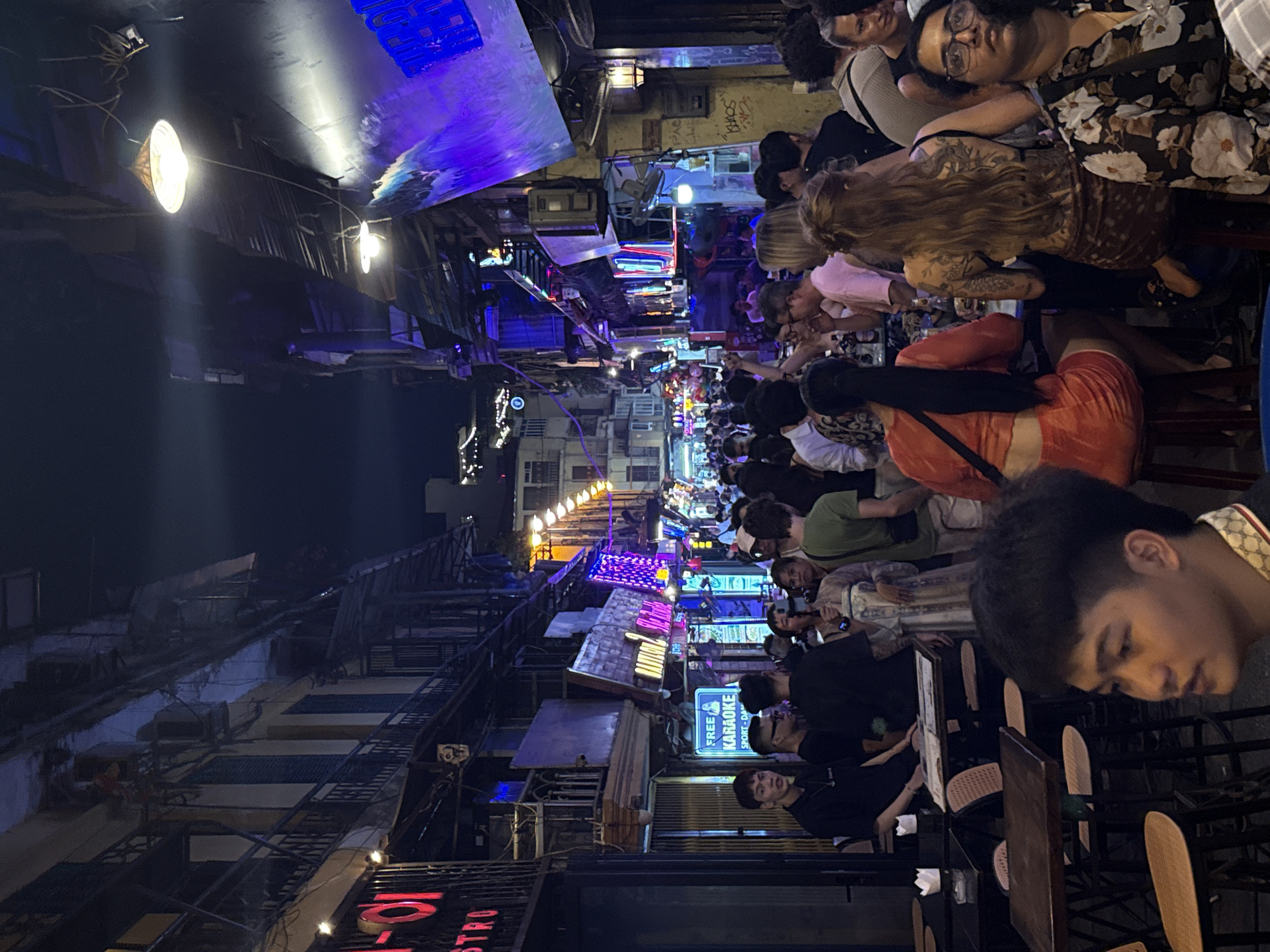 The image depicts a bustling street scene at night in Hà Nội, Vietnam. The street is narrow and crowded with people seated at tables and chairs that spill out from restaurants and bars. The buildings on either side are closely packed together, with a mix of architectural styles and visible utility infrastructure. Colorful neon signs and string lights illuminate the street, creating a vibrant atmosphere. Many of the people are interacting, eating, drinking, or simply observing the scene. A sign for "Free Karaoke" is visible. The overall impression is one of a lively and densely populated urban environment.