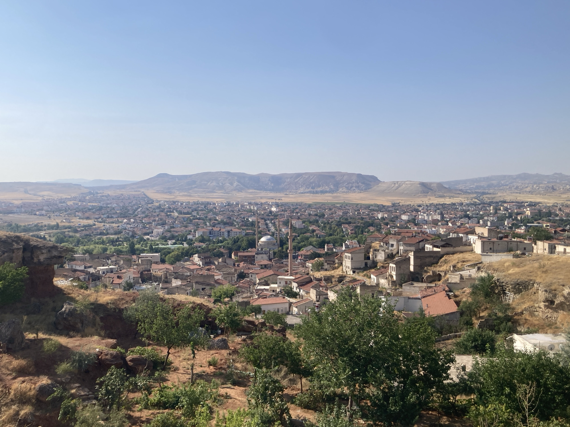 This is a high-angle, wide shot of the city of Avanos İlçe Merkezi, Turkey, under a clear, pale blue sky. The foreground is dominated by dry, rocky terrain with sparse green trees and shrubs. Behind this, the city sprawls across the landscape, characterized by densely packed buildings with red-tiled roofs. In the mid-ground, a mosque with a dome and two tall minarets stands out amongst the other structures. The city extends to the horizon, where a range of low, layered mountains is visible. The overall impression is of a sun-drenched, arid environment.