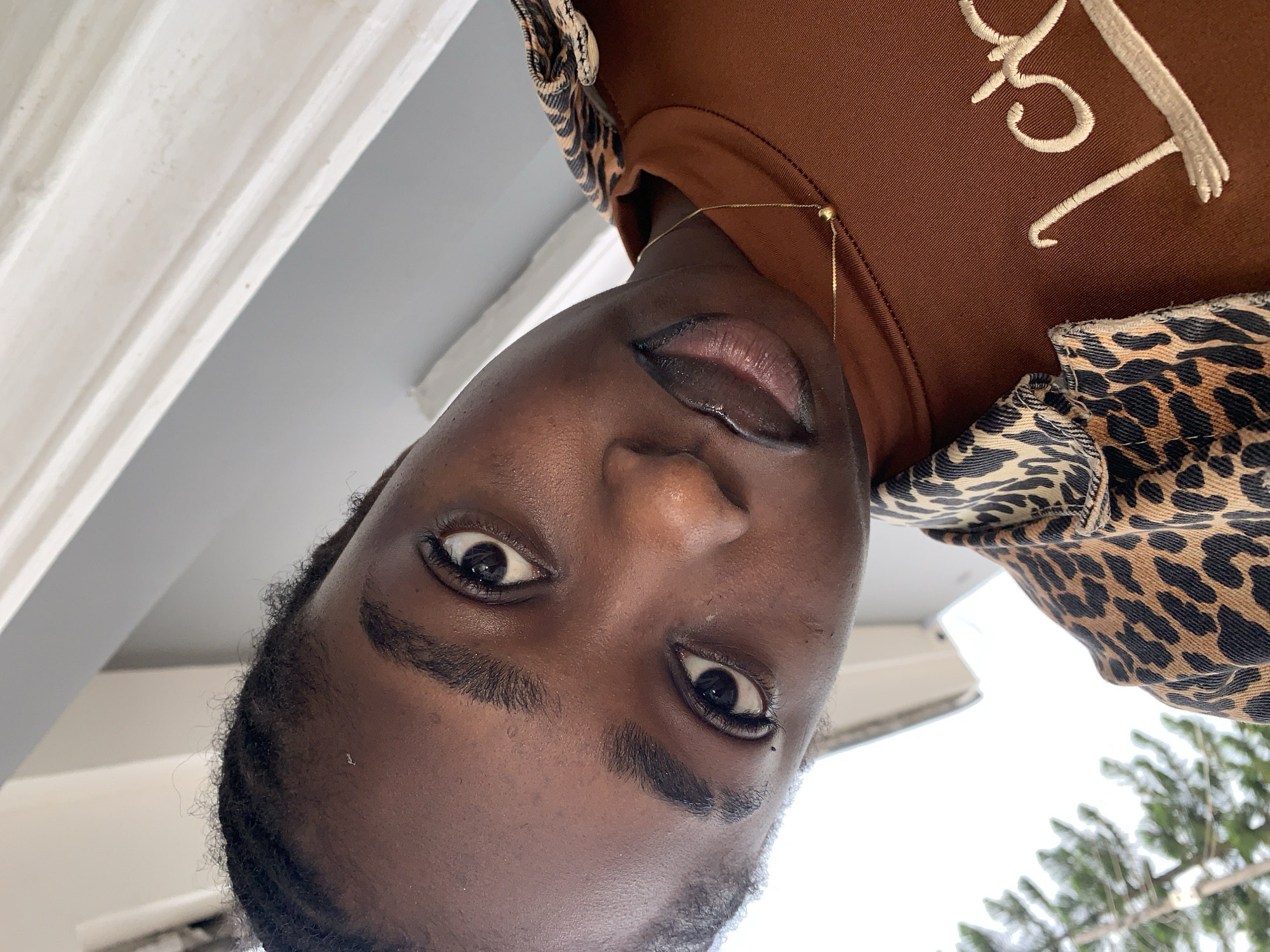 The image is a close-up, tilted selfie of a young Black person wearing a brown long-sleeved shirt with embroidered writing on the right side and a leopard-print jacket over it. They have braided hair, dark eyes, and are wearing subtle makeup, including eyeliner and lip gloss. A thin gold chain with a small ball pendant is visible around their neck. The background is blurred and appears to be outdoors with white trim and some greenery visible, suggesting a residential area. There is no specific indication of the location being Gura Top, Nigeria, as no recognizable landmarks or contextual cues point to that specific city.