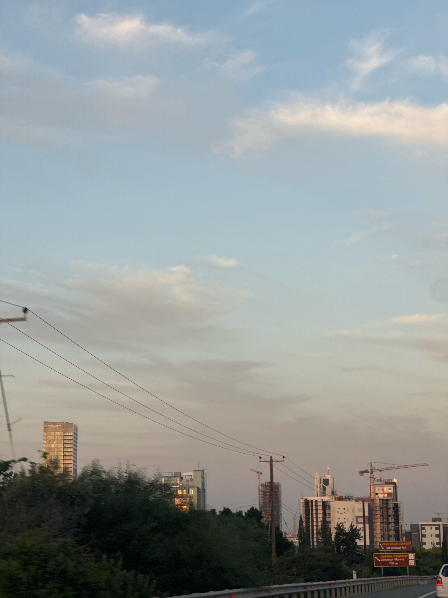 The image captures a view of urban development in the Agios Tychona Community, Cyprus, against a backdrop of a soft, cloud-filled sky. In the mid-ground, several modern buildings are visible, some appearing to be under construction, indicated by the presence of cranes. One prominent tall building displays "ATHANASIOU" on its facade. A series of utility poles with overhead wires traverse the scene from left to right. In the foreground, a highway barrier runs horizontally, suggesting the vantage point is from a road. The overall impression is one of a city undergoing growth and modernization, illuminated by the gentle light of dusk or dawn.