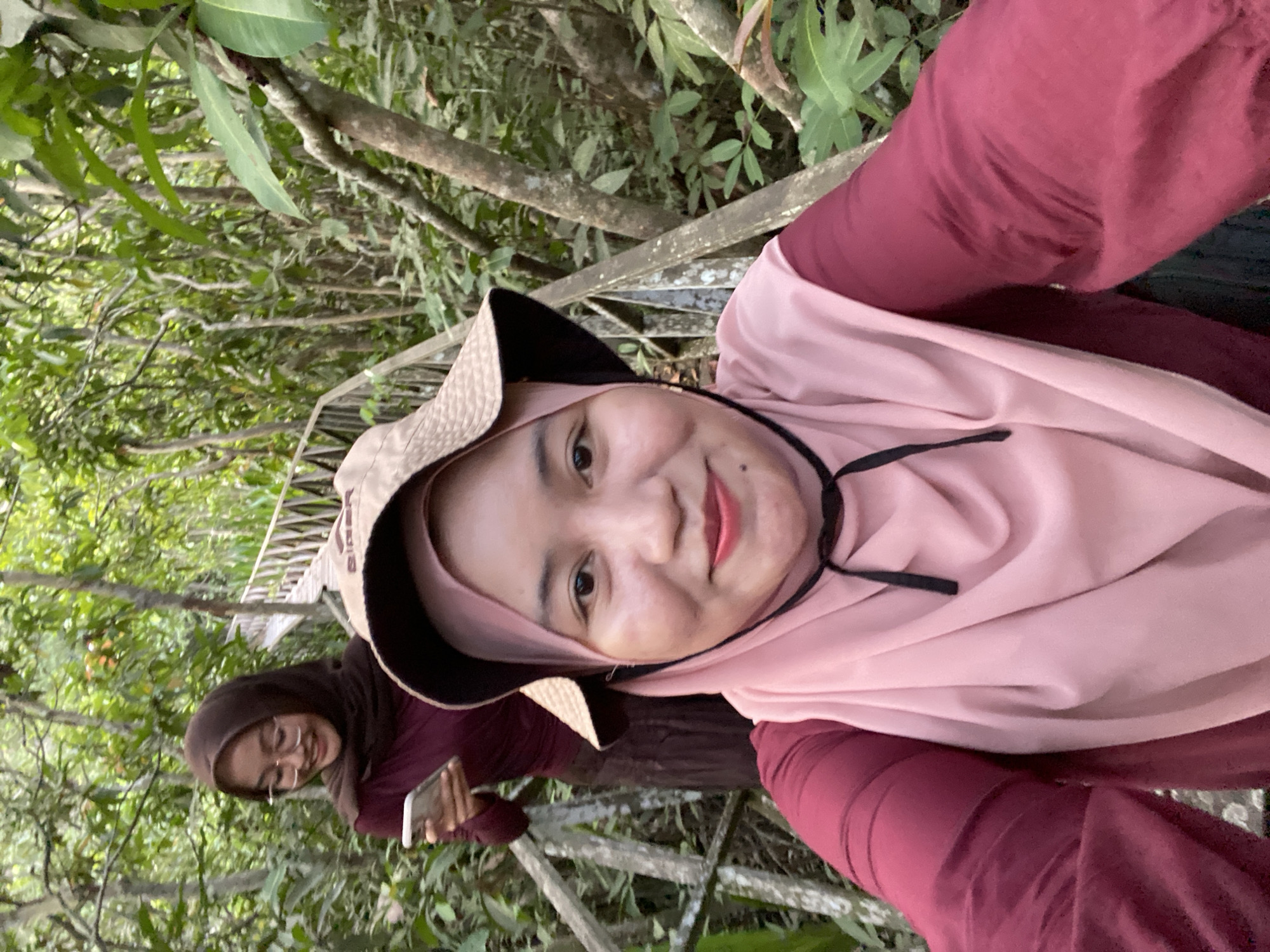 Two women are posing for a selfie on a wooden walkway through a lush, green forest. The woman in the foreground, wearing a maroon shirt and a light pink hijab with a wide-brimmed hat, is smiling at the camera. The second woman, in the background, is wearing a maroon long-sleeved top and a dark brown hijab, and she is looking down at her phone. The wooden walkway is constructed of weathered planks and railings and is surrounded by dense vegetation and tall trees. The overall impression is of a casual outing in a natural environment. The location is identified as Sungai Pitung, Indonesia.