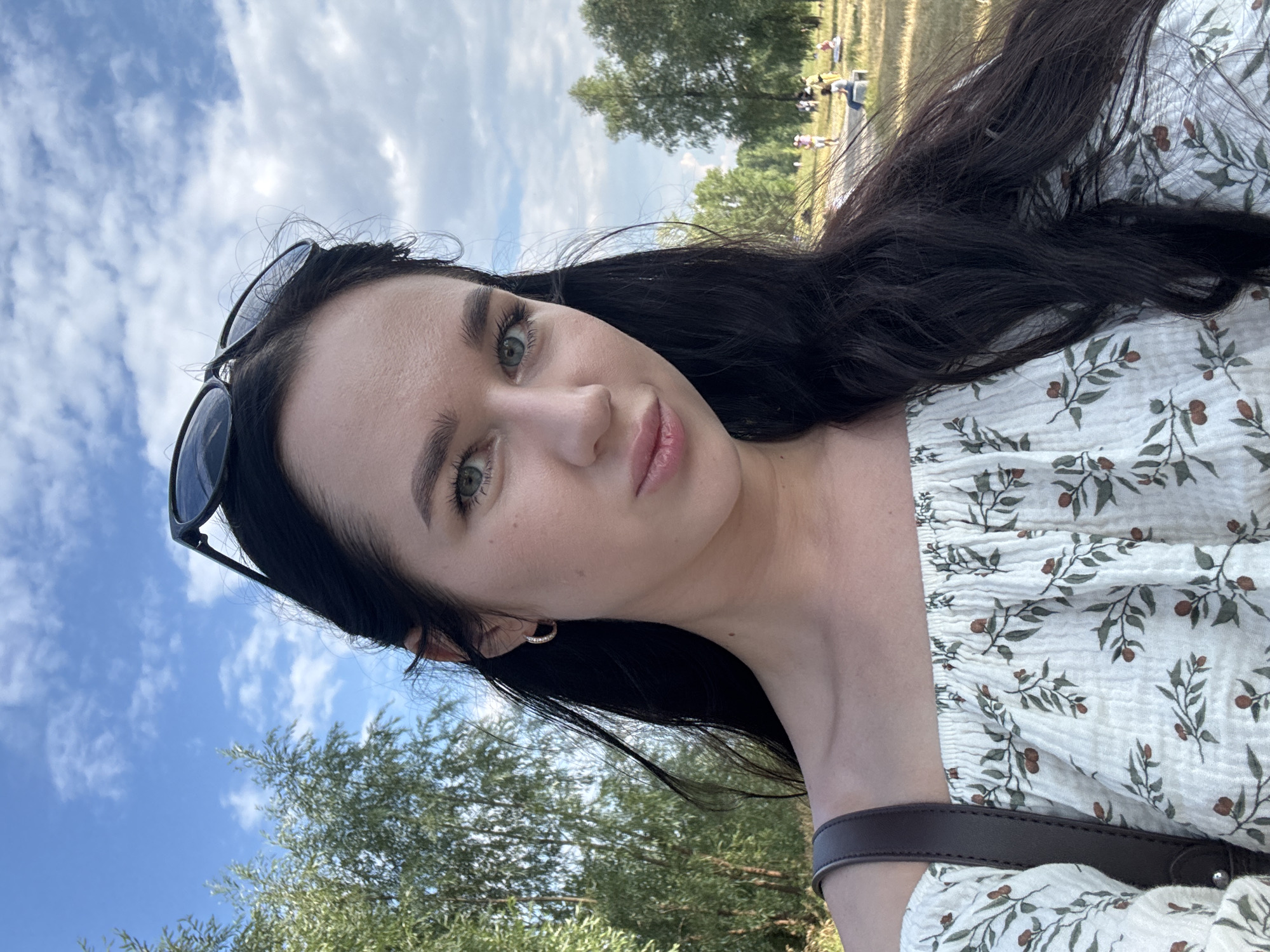 The image is a close-up selfie of a young woman with long, dark hair and blue-green eyes, wearing an off-the-shoulder white top with a floral pattern. She has sunglasses on her head and is wearing hoop earrings. She appears to be outdoors in a park-like setting, with trees and a cloudy blue sky in the background. In the distance, several people are visible sitting and standing on a grassy area. The overall impression is of a casual, sunny day. The location is not specified in the image, and therefore cannot be identified as Novosibirsk, Russia.
