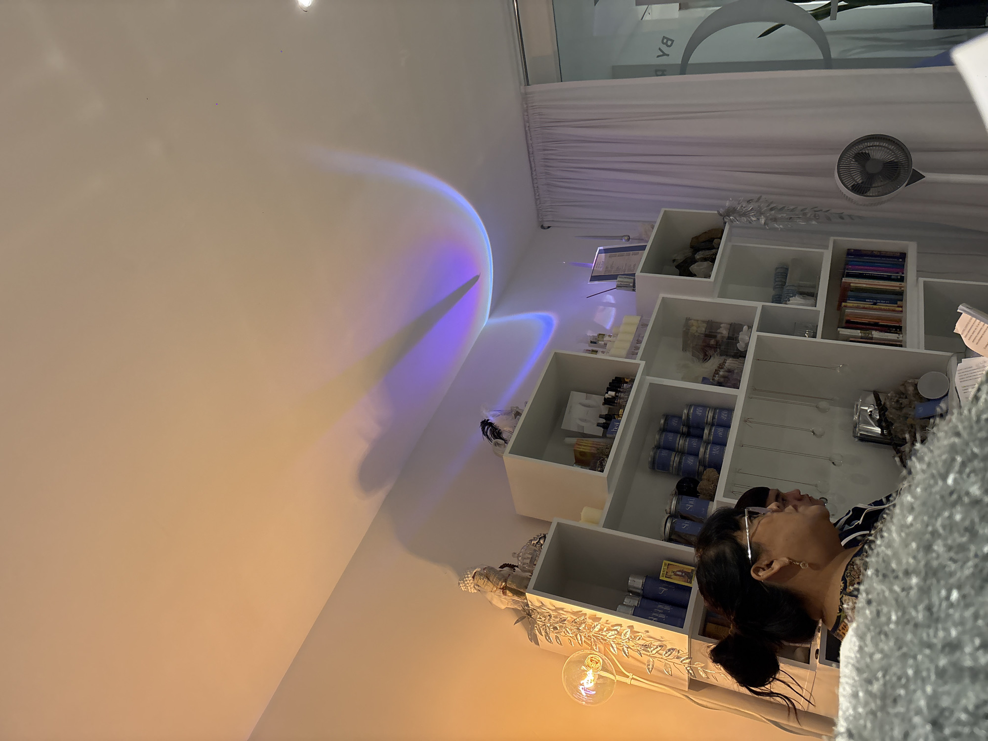 A woman wearing glasses and a bun hairstyle is captured in an indoor shot with shelves of various items in the background. The lighting is subdued, casting shadows across the room, and a blue arc is projected on the wall. A white curtain and a small fan are also visible in the background. There is no visible information to confirm the location in the city of Puebla, Mexico.