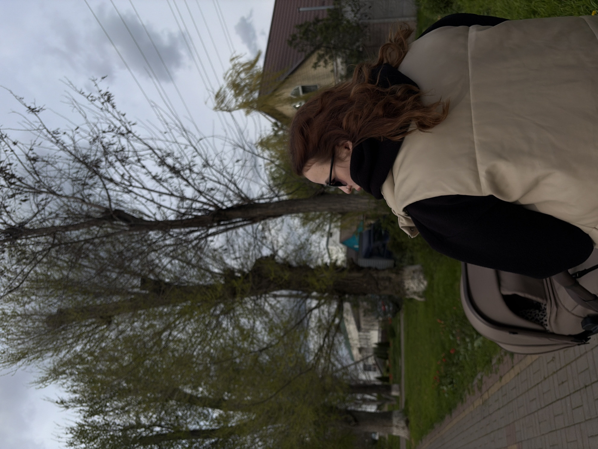 A slightly blurred, sideways photograph shows a woman pushing a stroller on a sidewalk in Novopokrovskaya, Russia. The woman has reddish-brown curly hair and is wearing glasses, a black hooded top, and a cream-colored puffy vest. The stroller is a muted gray color. To the left of the sidewalk are several trees with sparse leaves, with buildings visible between them. Overhead are power lines against a cloudy sky. The sidewalk is made of gray bricks.
