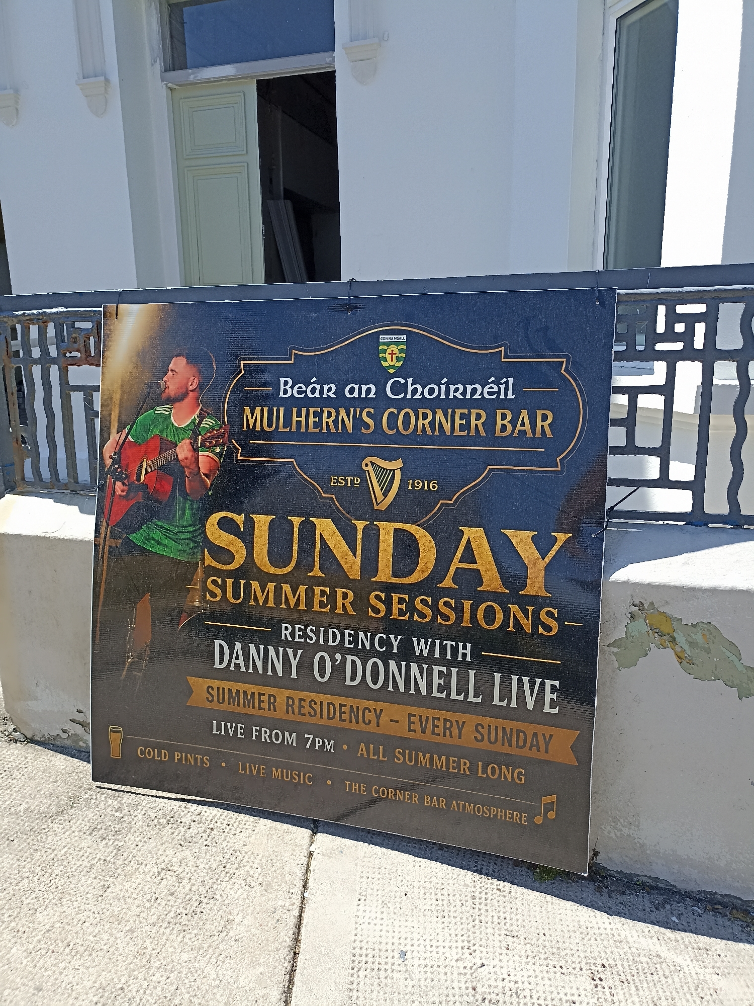 A sign for Mulhern's Corner Bar is displayed outside a building. The sign advertises "Sunday Summer Sessions" with "Danny O'Donnell Live" as a residency every Sunday from 7 PM, "all summer long." It also mentions "cold pints, live music, and the corner bar atmosphere." The sign features a graphic of a man playing an acoustic guitar and a harp emblem. The building appears to be a white-painted structure with windows. The ground in front of the sign is paved with light-colored stones. A metal railing is visible behind the sign.