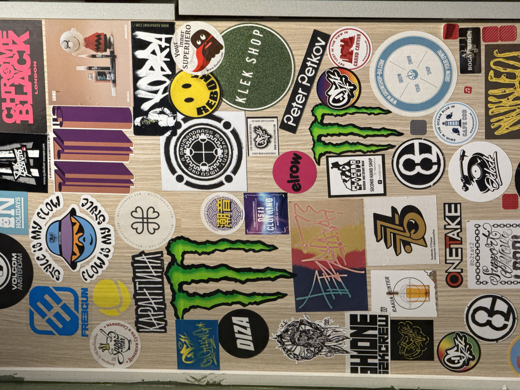 The image is a close-up overhead shot of a flat surface covered in stickers, arranged in a dense, overlapping pattern.
Many stickers show brand logos or imagery associated with street culture and skateboarding. Noteworthy brands include Volcom, Freegun, Monster Energy, and others.
Several stickers feature text, some in Cyrillic script indicating the presence of brands, locations, or themes related to Bulgaria. One sticker explicitly mentions "Sofia," the capital city of Bulgaria. "Str. Oborishte 80 Sofia" points to a specific street and address within the city.
Other stickers show abstract art, geometric designs, and stylized typography. There are round stickers with logos and imagery related to music or events. Some stickers are related to clothing brands and shops.