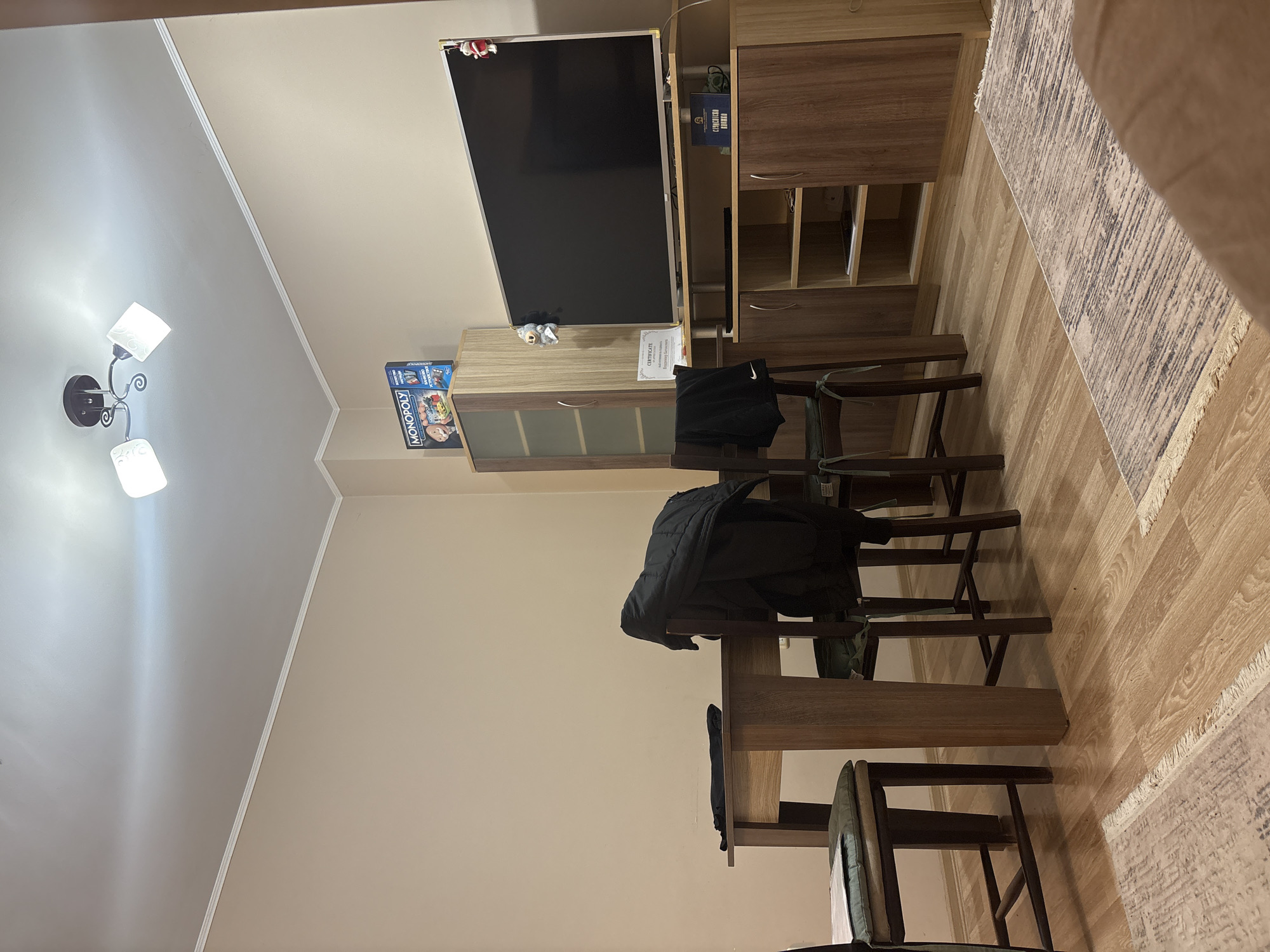 The image captures an indoor scene, likely a living room or dining area, furnished with modern wooden pieces. The perspective is from one end of the room, looking towards a media console and dining table.
Key subjects include a large flat-screen television mounted above a wooden media console, which features both open shelves and closed cabinets. To the left of the main console, there's a smaller cabinet unit with a clear glass-fronted section. A dark wooden dining table occupies the foreground and midground, surrounded by four matching wooden chairs.
No people are visible in the scene. The room appears static, showcasing its interior design and furnishings.
Notable details include the room's color scheme: light beige walls are topped with white ceiling molding, leading to a white ceiling. An overhead chandelier with three light fixtures provides illumination, with at least two bulbs visibly lit. The floor is covered with light-colored wooden or laminate planks, partially covered by a light-toned, patterned rug in the foreground.
Several objects are placed around the room:
- A "MONOPOLY" board game box is visible on the smaller cabinet unit.
- A white document, identified by the prominent text "CERTIFICATE" at the top, stands upright on the same cabinet.
- A dark jacket, possibly black, is draped over one of the chairs at the dining table.
- Another dark item, appearing to be a hooded sweatshirt with a white Nike swoosh logo, is draped over a chair closer to the TV unit.
- On a shelf within the main media console, a book with the partial title "COMMANDO" is visible.
The lighting suggests an indoor setting where artificial light is in use, potentially evening or an overcast day. No outdoor views or specific location cues beyond the furnishings are visible in the image itself.